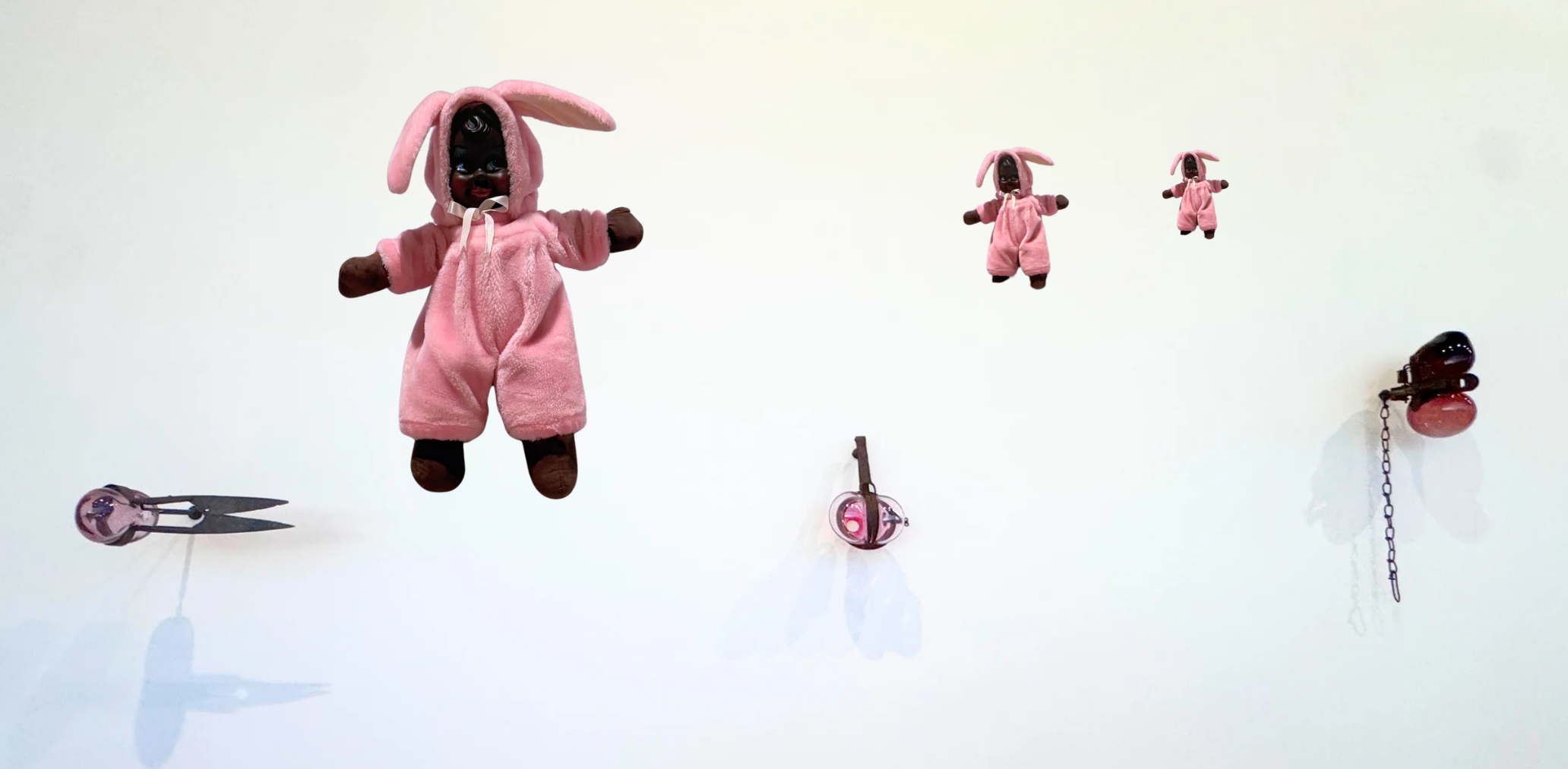 Three plush dolls dressed in pink bunny costumes, floating against a white background. There are also various small objects, including a pair of scissors and small round items attached to chains, around them.