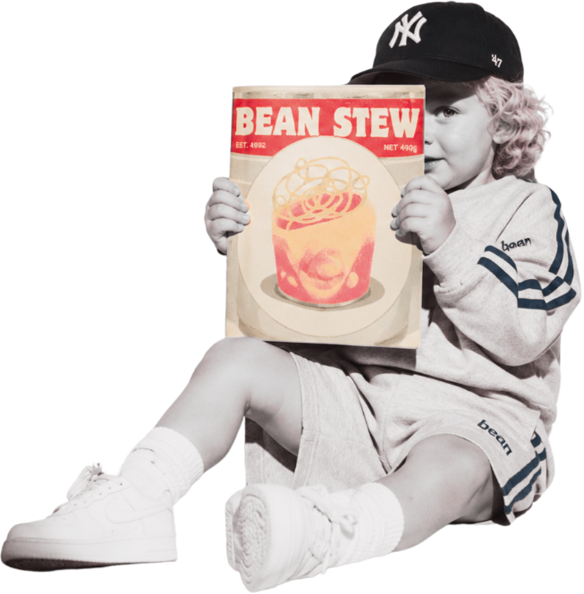 A young child sitting down wearing a baseball cap, sweatshirt, shorts, and sneakers, holding a box of Bean Stew with an illustrated face and noodle design on the front.