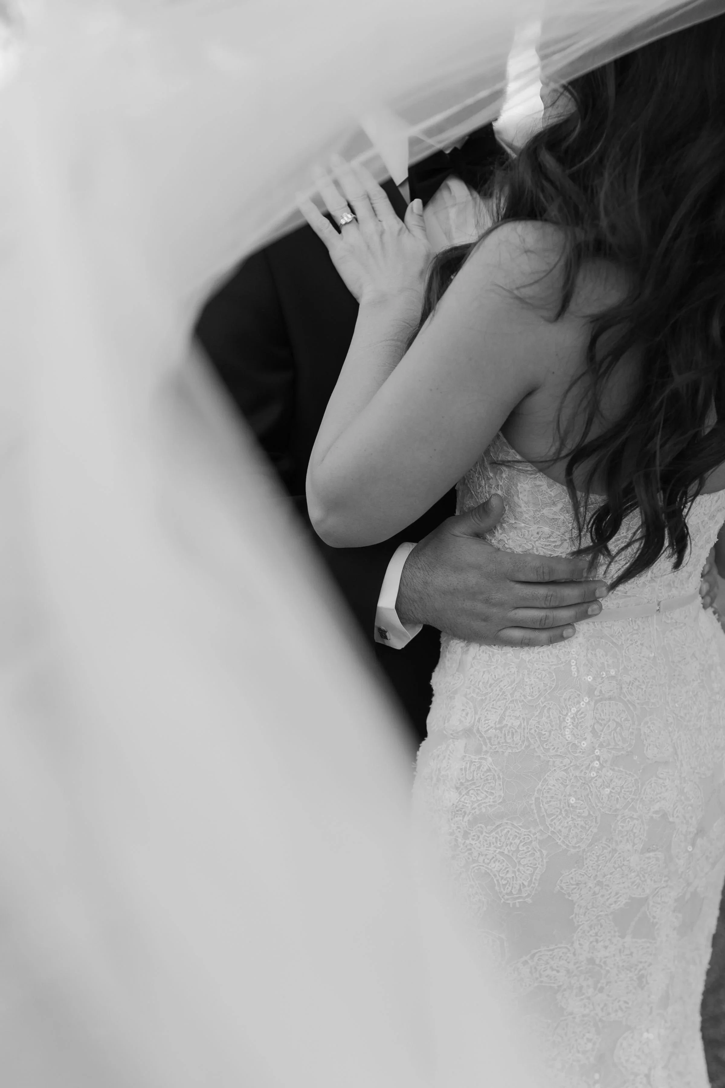 Black and white photo of a bride and groom, with the bride leaning into the groom, her hand on his chest, and the groom holding her waist, capturing an intimate moment.