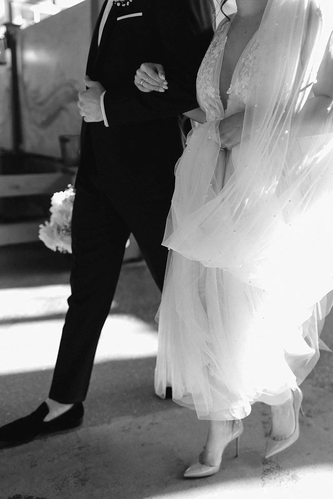 A black and white photo of a bride and groom standing together inside. The bride is wearing a lace wedding dress with a veil and high heels, while the groom is in a dark suit and tie, holding a book or folder.