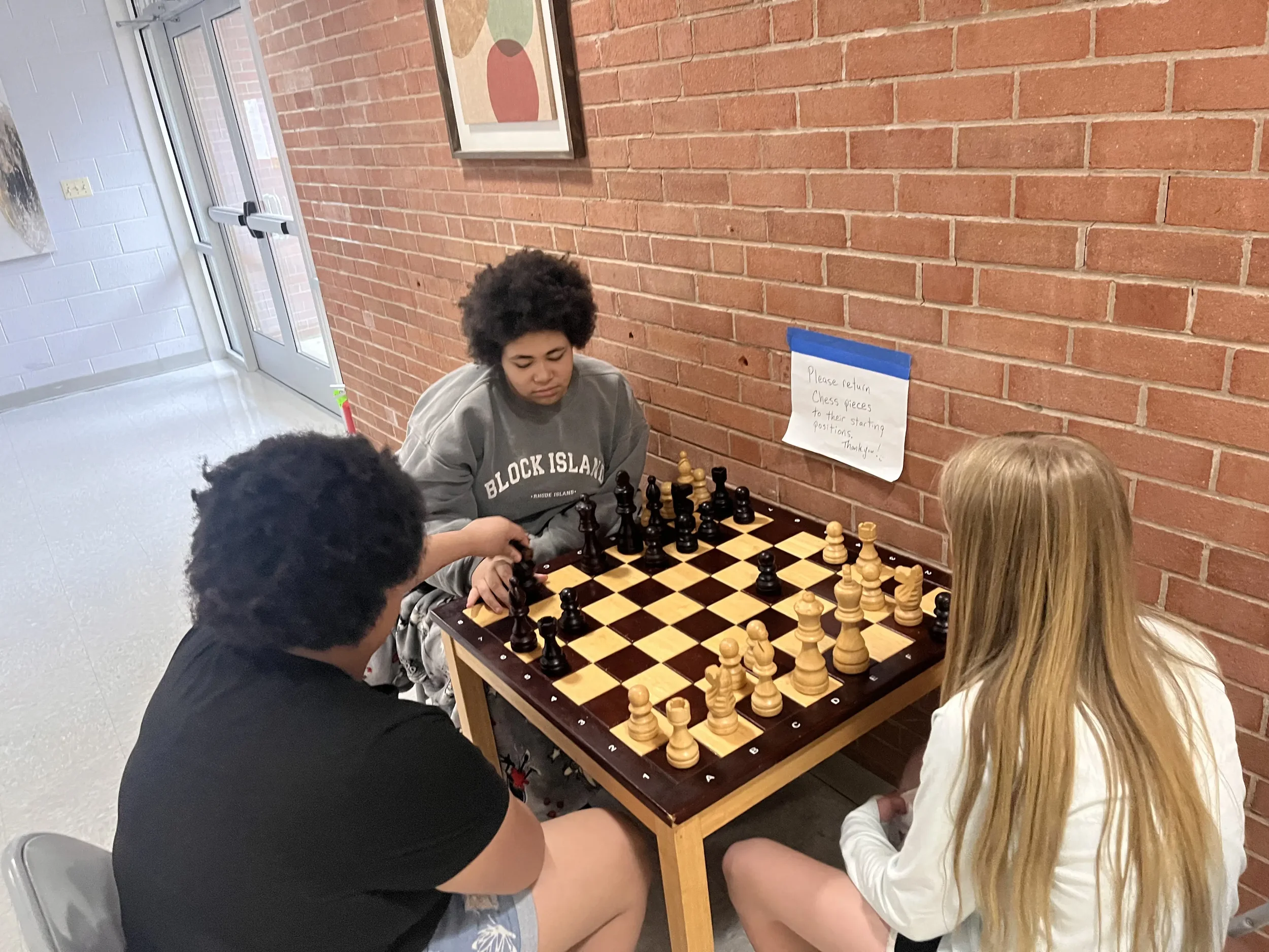 Young people playing chess at a table against a brick wall with a framed picture hanging on it and a handwritten note on blue tape.