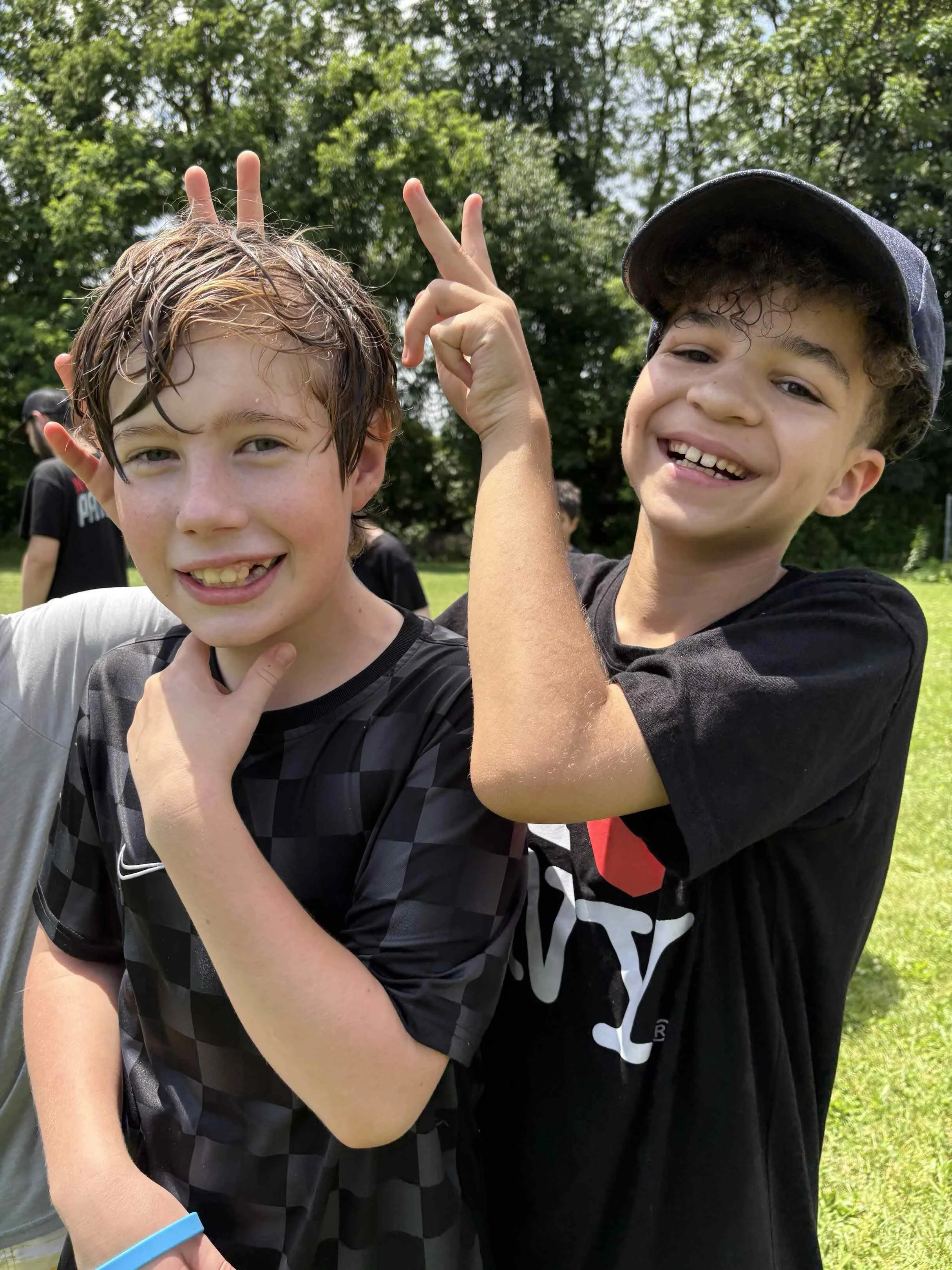 Two smiling boys outdoors making peace signs with their fingers. They are wearing casual T-shirts, and the background shows green trees and a grassy area.