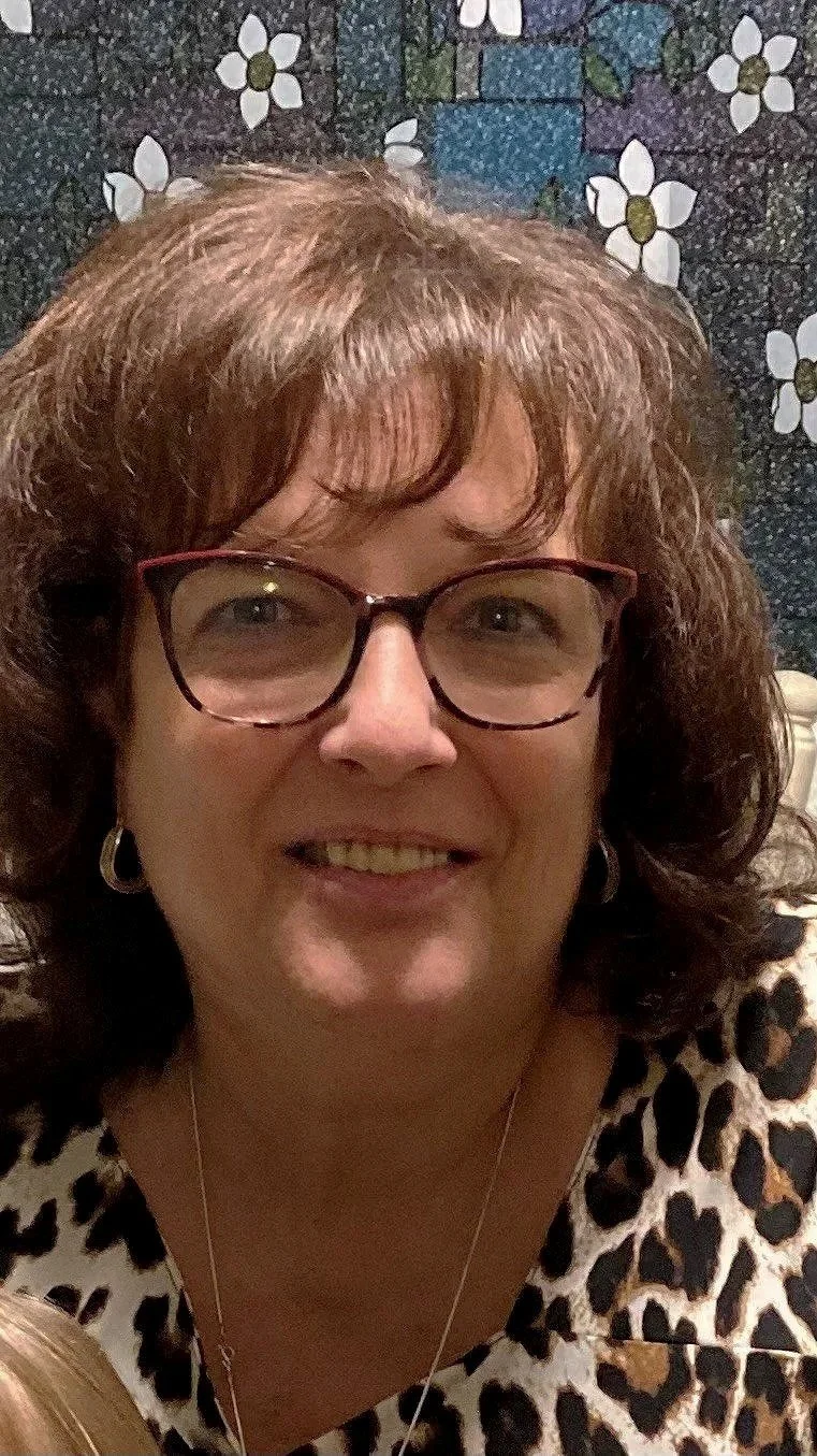 A woman with glasses and a bun hairstyle standing outdoors on a sunny day, wearing a cream-colored sweatshirt with U.S. Irish Football design and a necklace with a cross.