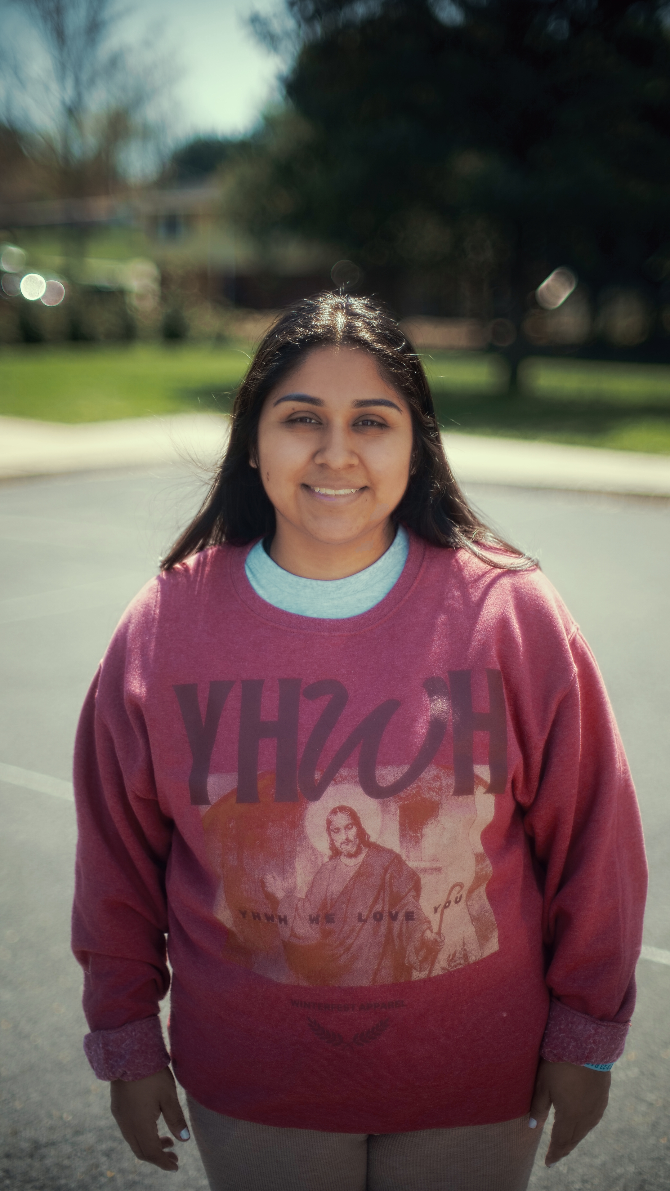 A woman smiling outdoors on a sunny day, wearing a vintage band-style sweatshirt featuring an image of Jesus Christ. She has long dark hair and stands in a parking lot with trees and buildings in the background.