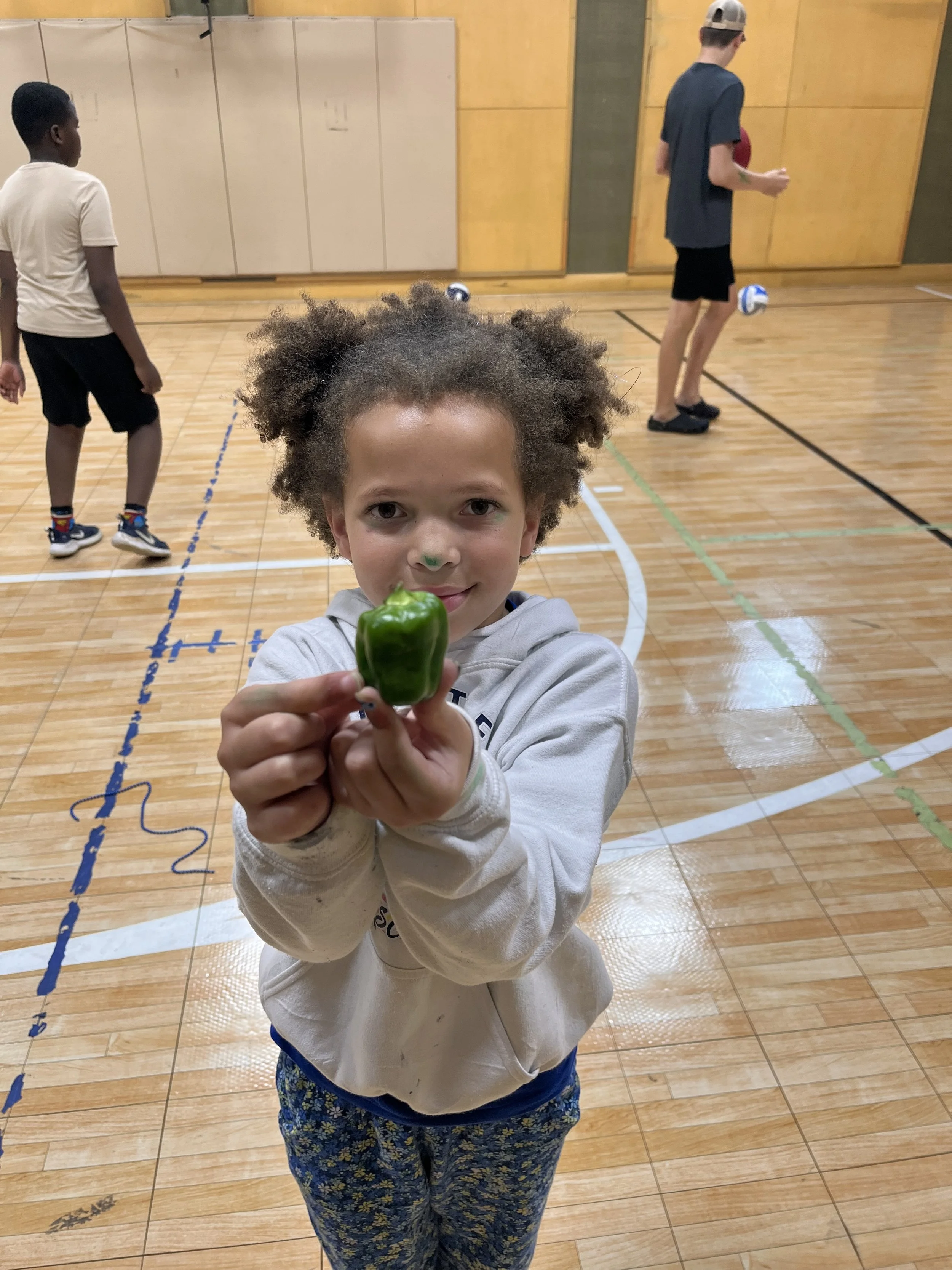 A young girl with curly hair and a gray hoodie holding a green bell pepper in a gymnasium. In the background, a boy is playing volleyball, and another person is playing volleyball on the court.