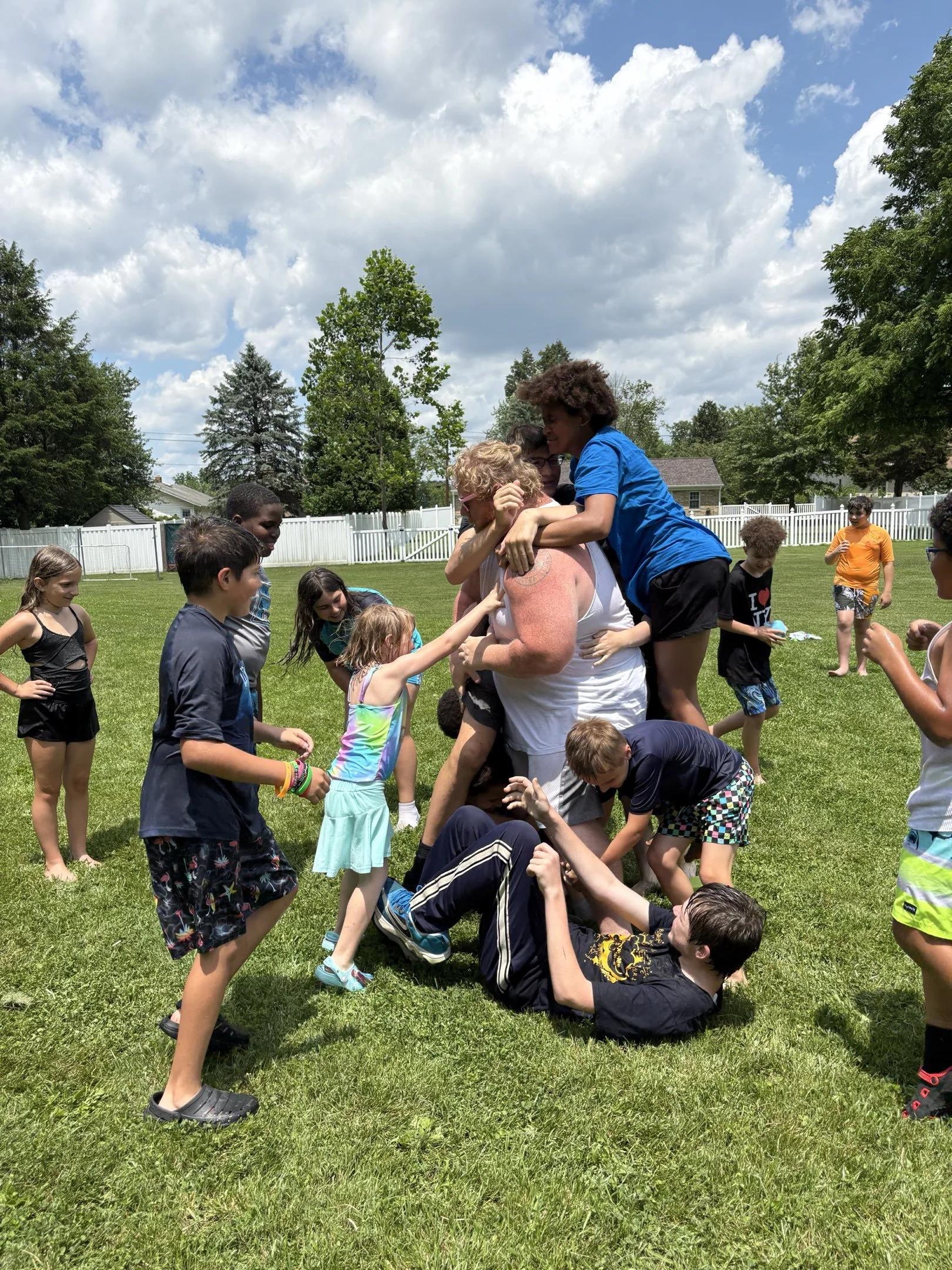 Children and teenagers playing a game of human foosball outdoors on a sunny day with a white fence, green trees, and houses in the background.