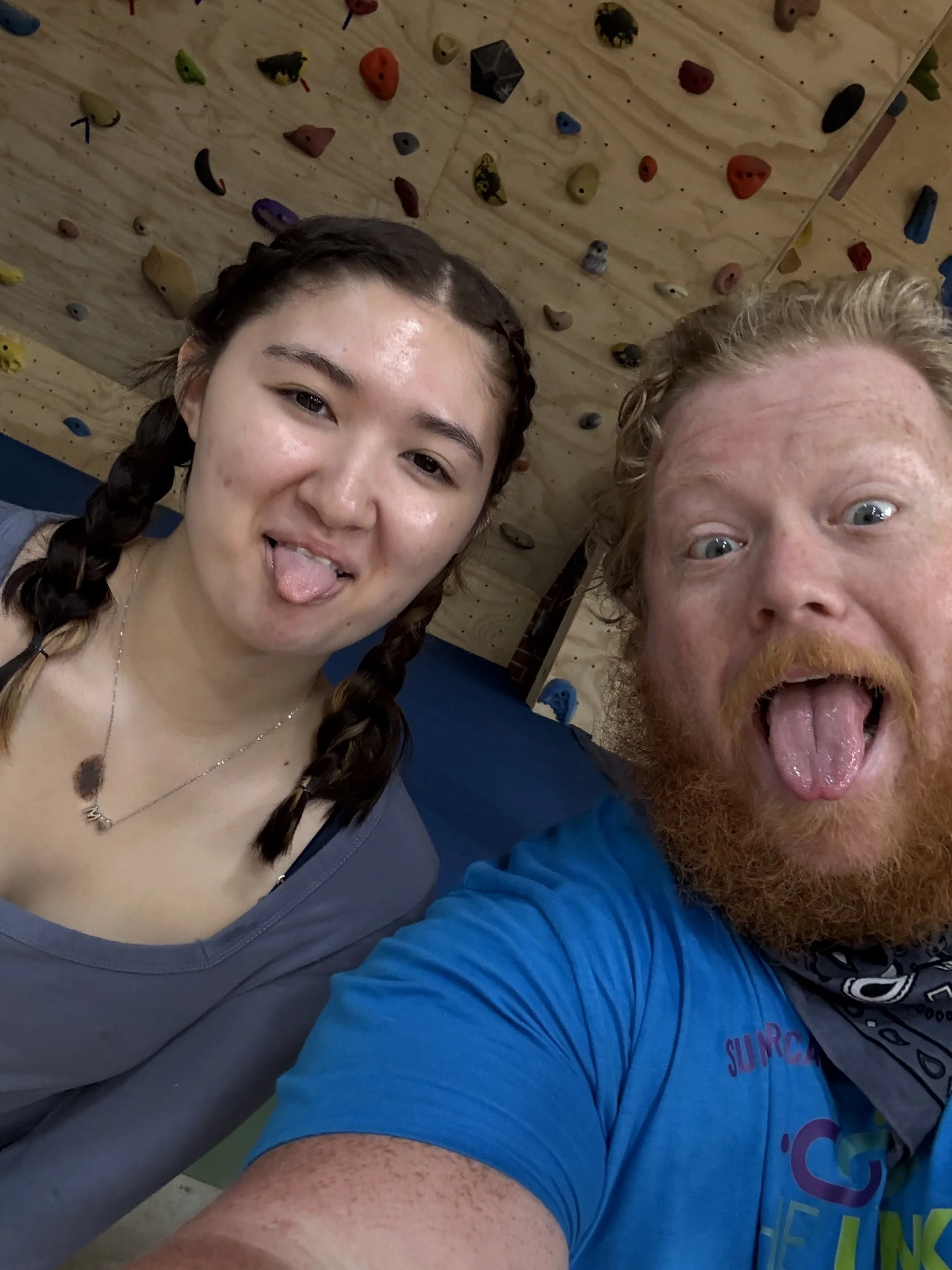 Two people taking a selfie in front of a rock climbing wall. The woman on the left has dark braided hair, sticking out her tongue, and wearing a gray shirt. The man on the right has curly blond hair, a beard, and is also sticking out his tongue, wearing a blue shirt. The climbing wall behind them has various colorful holds.
