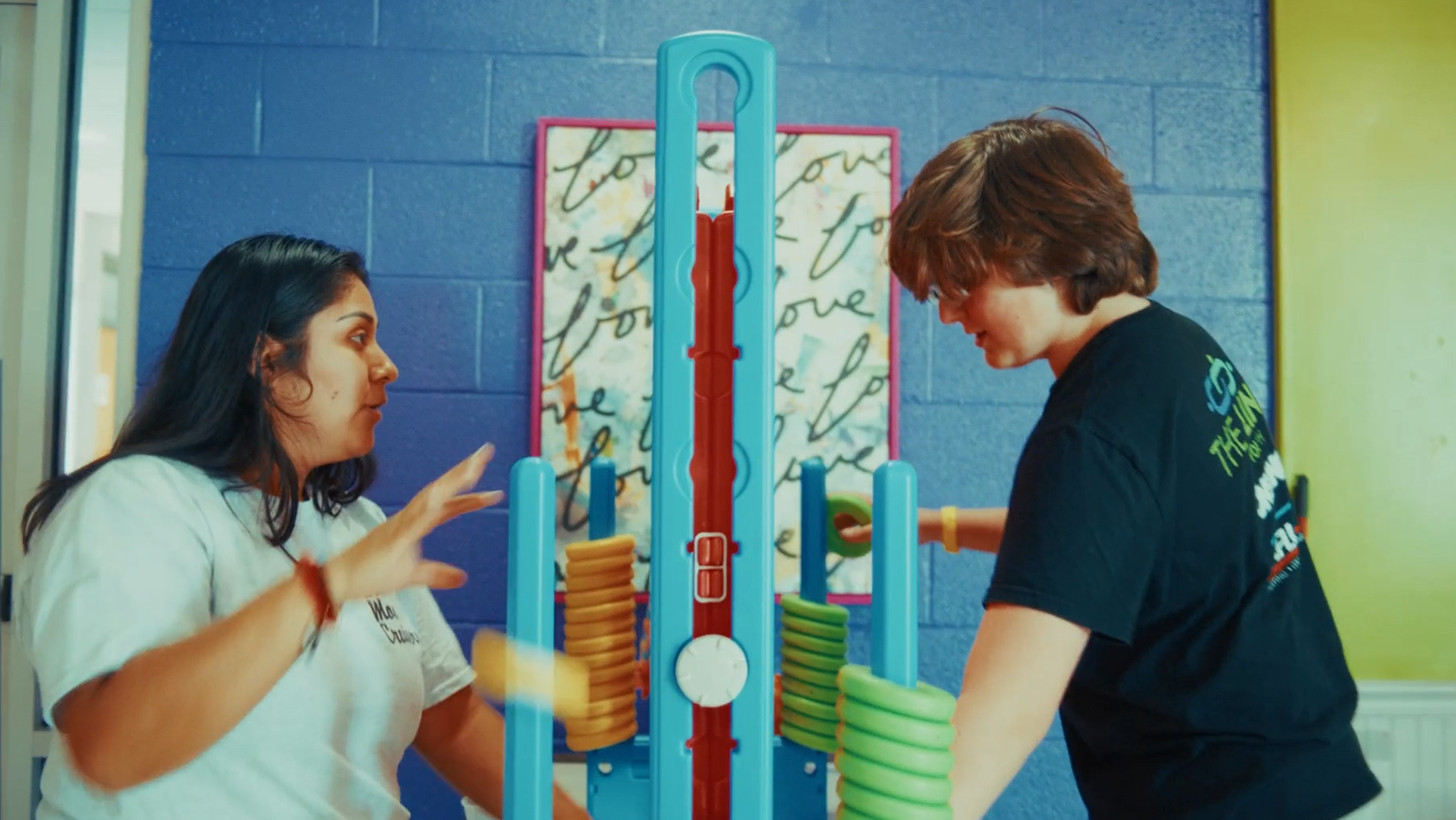 Two people playing a giant ring toss game indoors. A woman with dark hair on the left and a young man with brown hair on the right face each other, focusing on the game. The background shows a blue wall with a decorative art piece.