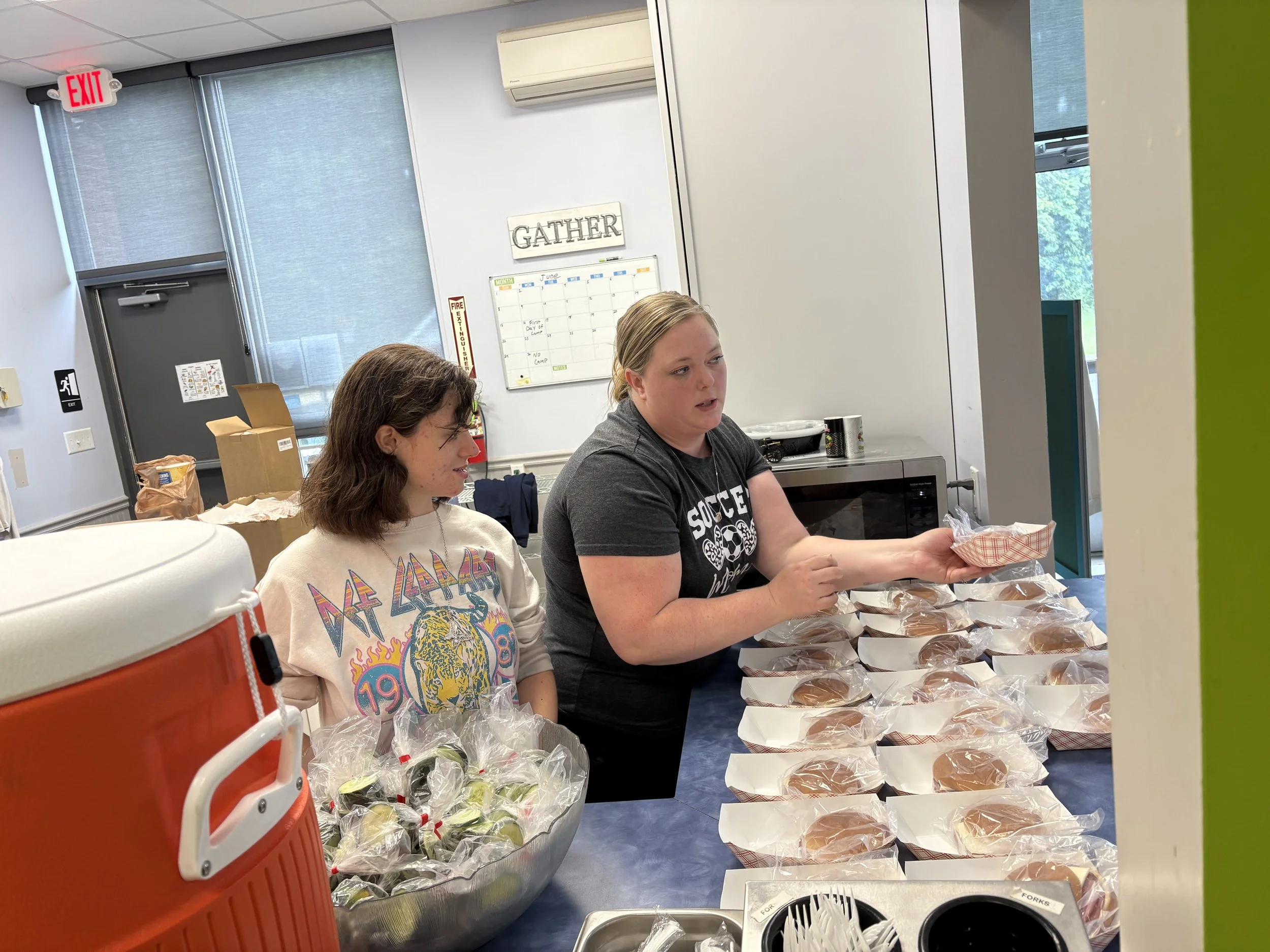 Two women are sitting at a table with pre-packed sandwiches and salads, in a room with windows, a whiteboard, and an air conditioner, possibly in a community center or office.