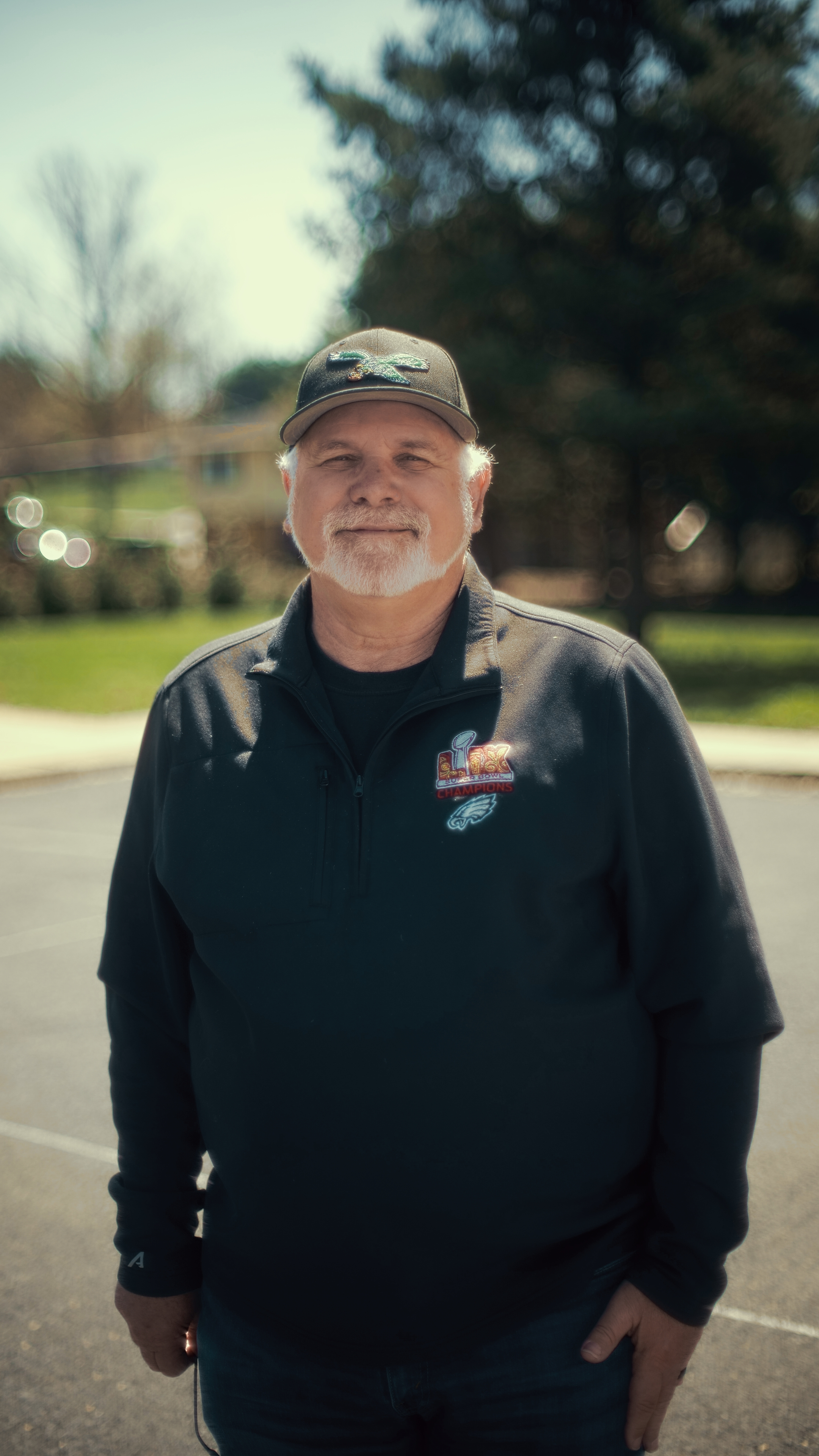 A smiling man with a gray beard and mustache wearing a black jacket with a patch, a gray cap with a crocodile logo, standing outdoors in a parking lot with trees and houses in the background.