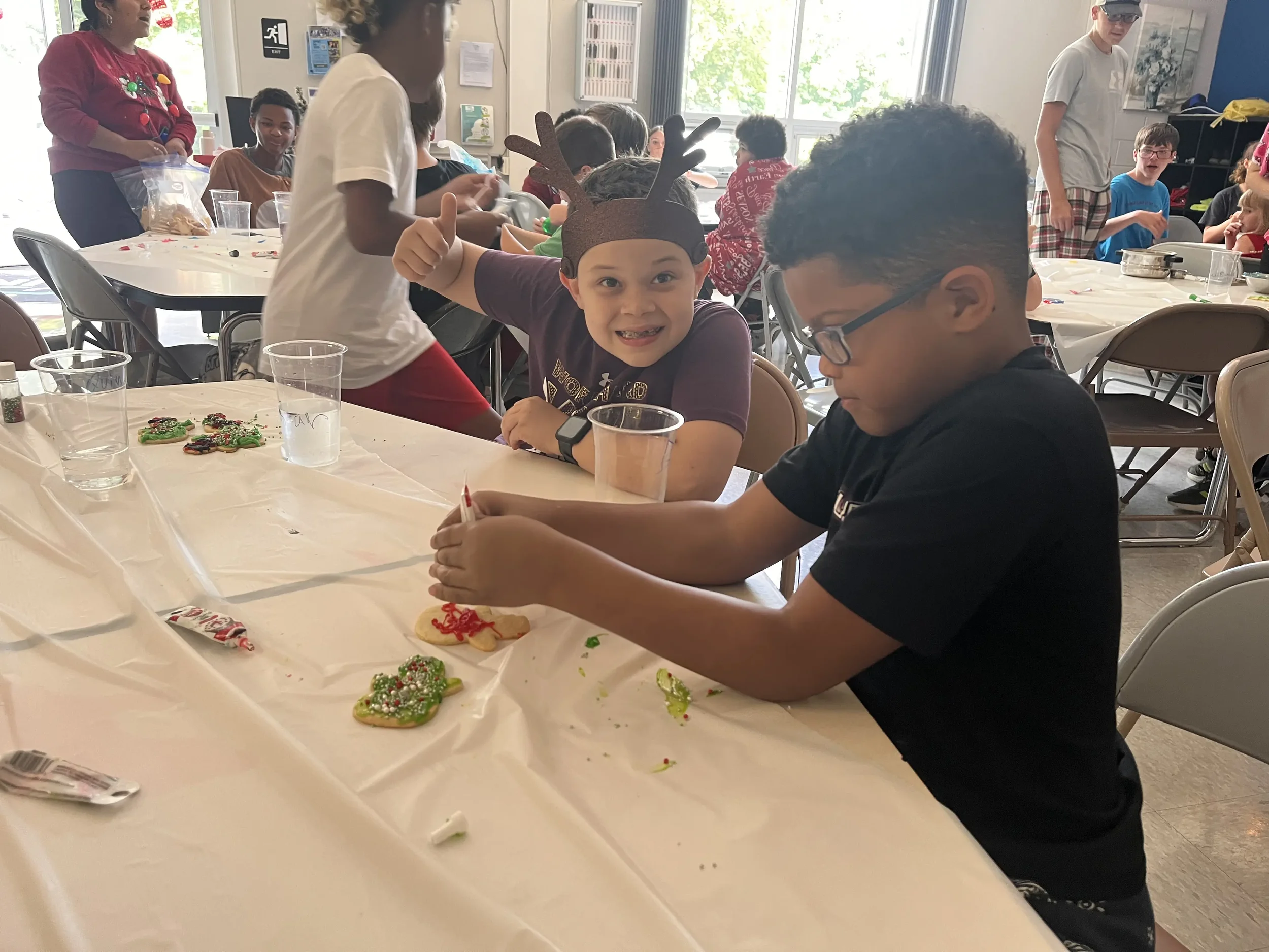 Children and adults in a room decorated for Christmas, making and decorating Christmas cookies with icing and sprinkles on a long table covered with a white tablecloth. Some children wear reindeer antler headbands.