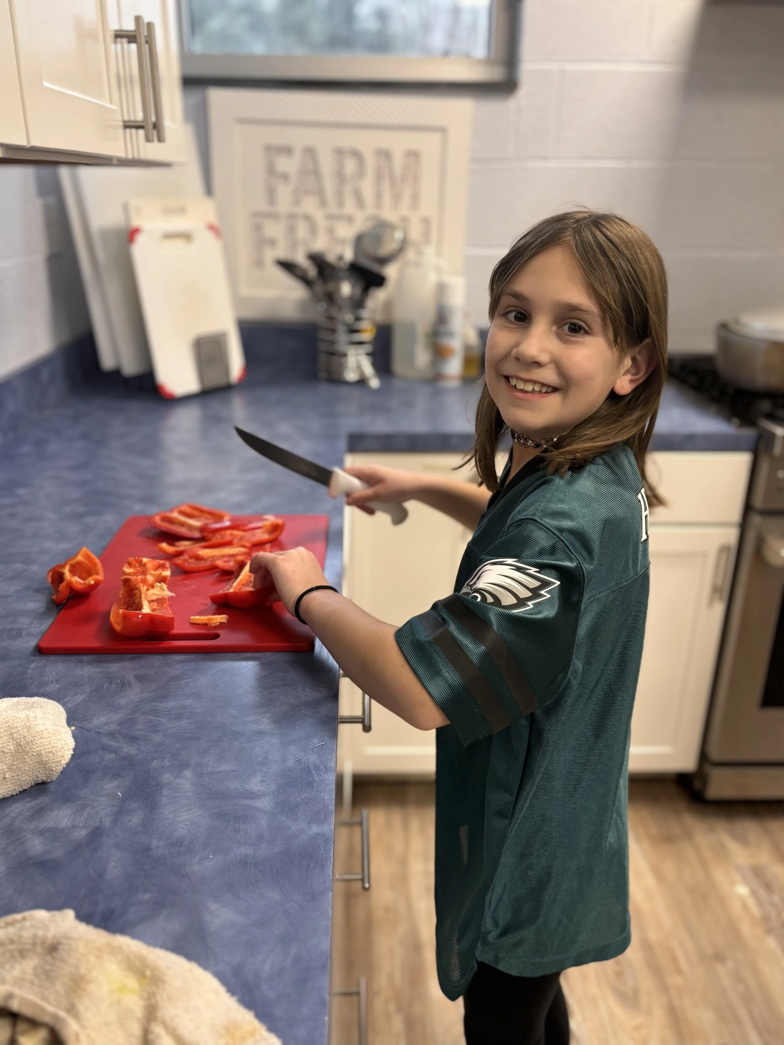 A young girl with brown hair smiling while chopping red bell peppers on a red cutting board in a kitchen.