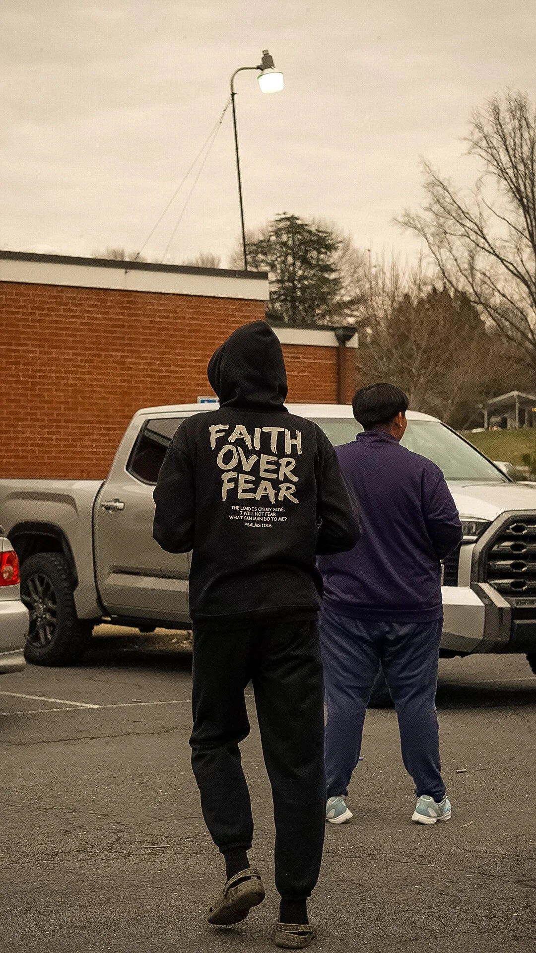 Two people walking in a parking lot, one wearing a hoodie with 'Faith Over Fear' written on the back, and the other in a purple jacket. There are parked cars and a brick building in the background.