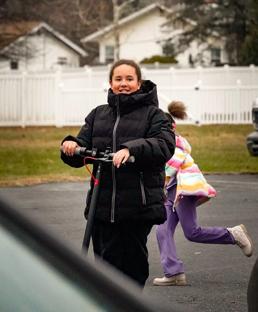 A smiling young girl in a black winter coat riding a scooter on a driveway, with another child in a colorful coat and purple pants running in the background.