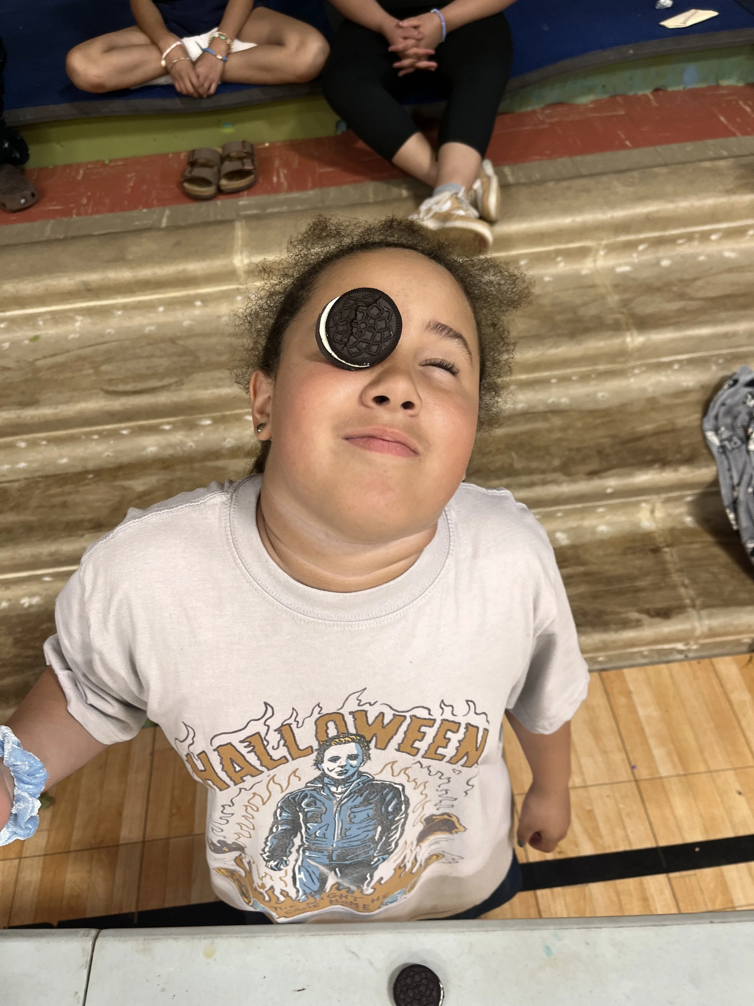 A young woman with curly hair lying on a wooden floor, with an Oreo cookie on her left eye. She is wearing a white Halloween-themed T-shirt with a skeleton graphic and the word 'HALLOWEEN'. In the background, there are other individuals sitting cross-legged on a mat, with shoes placed on the floor near them.