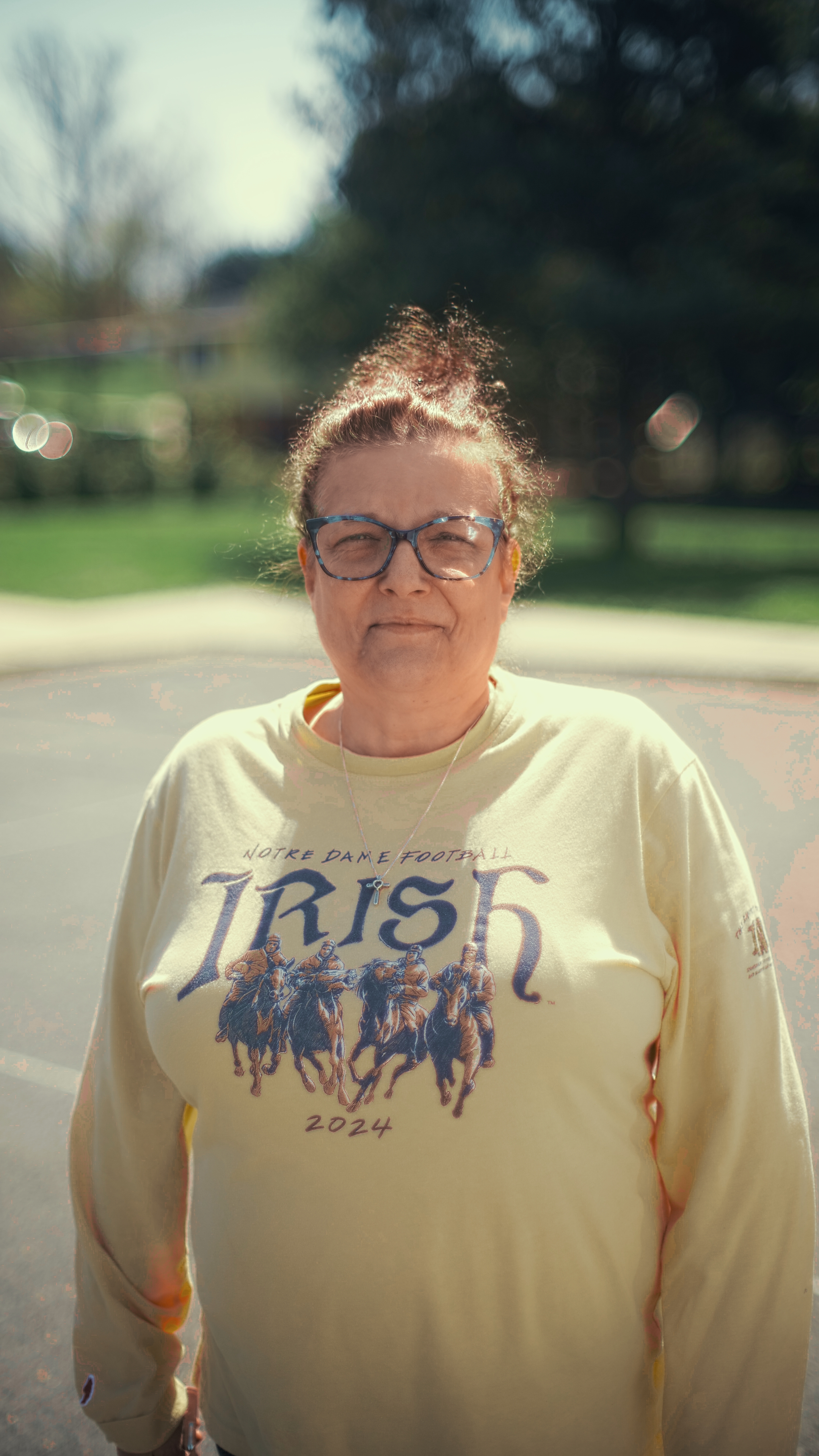 A woman with glasses and a bun hairstyle standing outdoors on a sunny day, wearing a cream-colored sweatshirt with U.S. Irish Football design and a necklace with a cross.