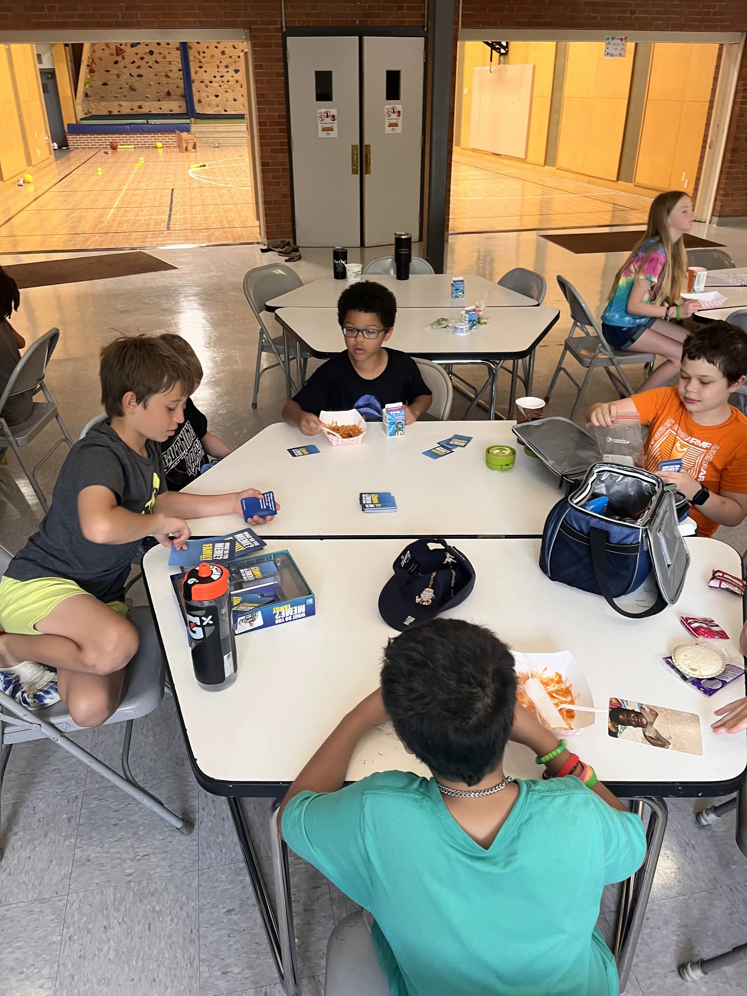 Children sitting around a table playing card games and eating snacks, with a gymnasium visible in the background.