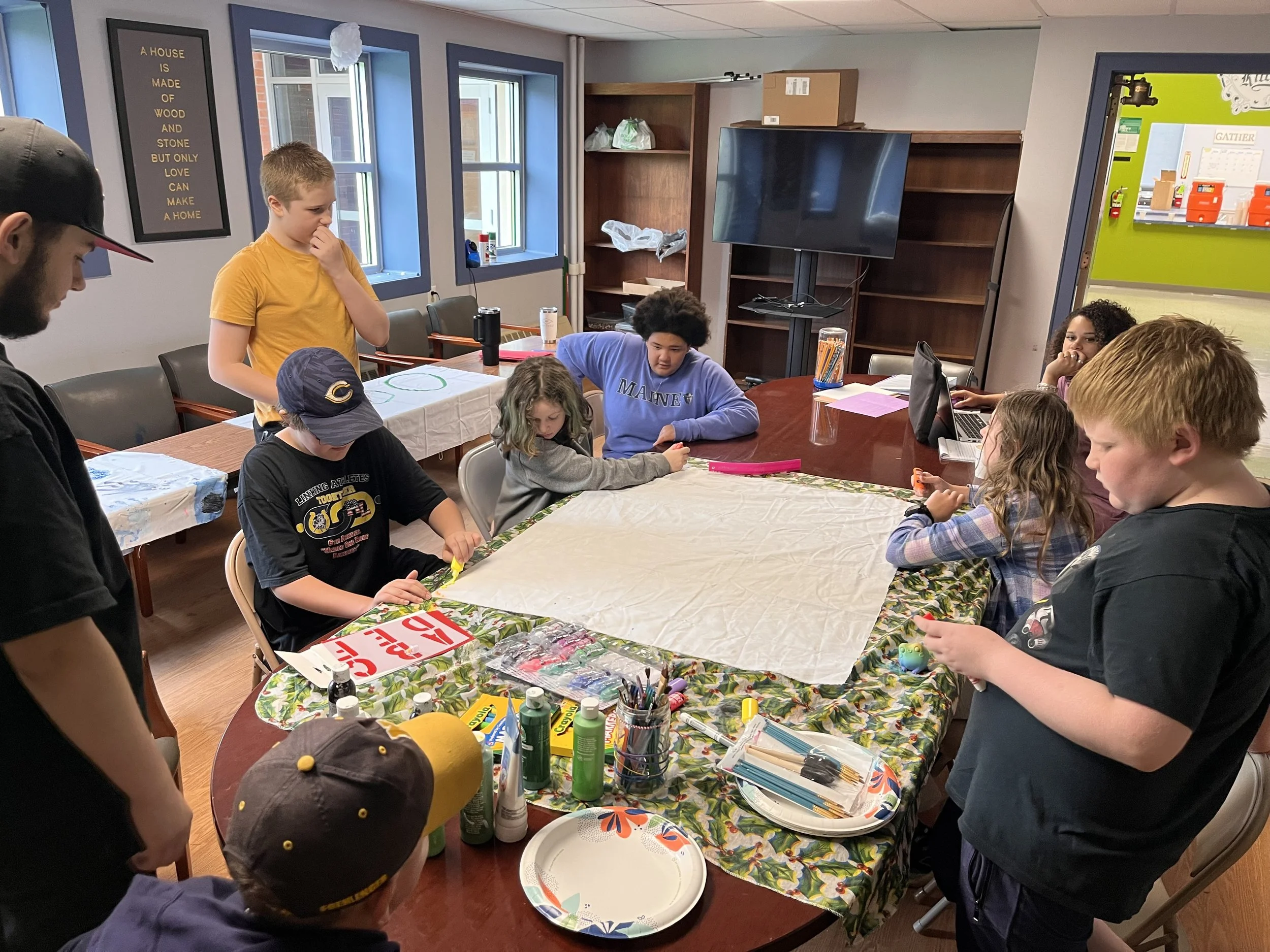Group of children and adults gathered around a table decorated with a festive tablecloth, engaging in arts and crafts activities in a community room with windows, bookshelves, and a large TV mounted on the wall.