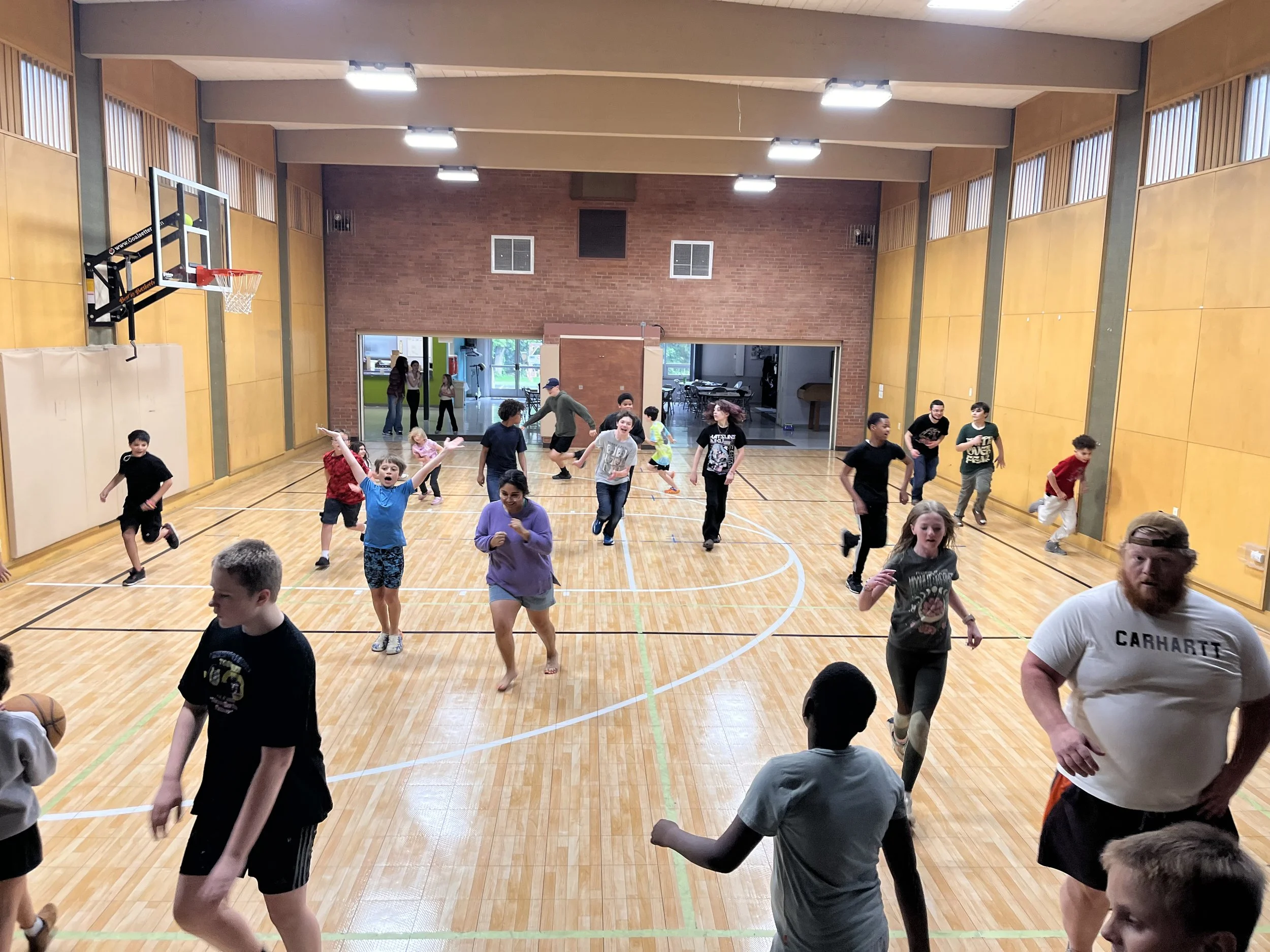 Children and teenagers playing and running in a gymnasium with a basketball hoop and basketball.