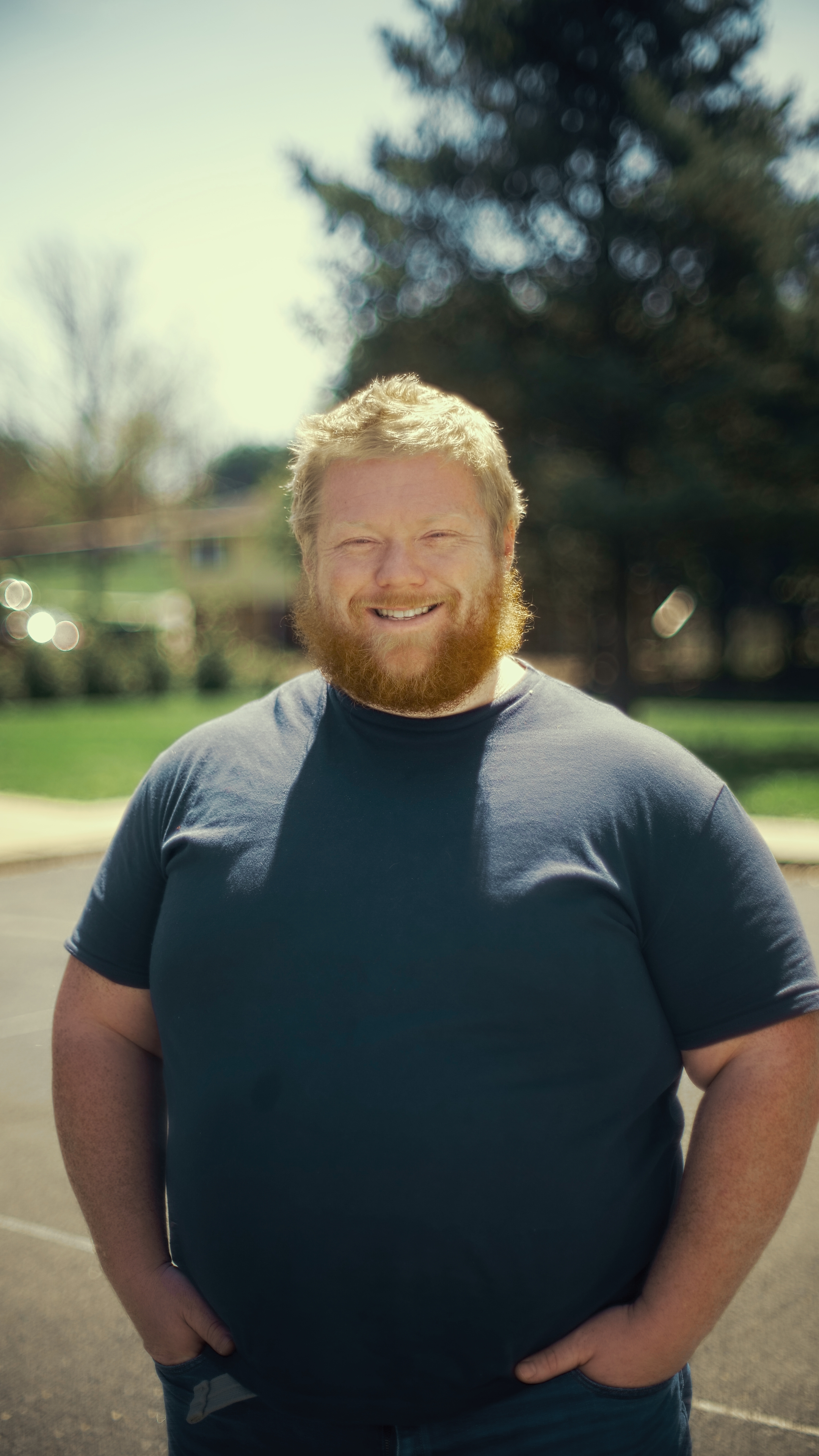 A smiling man with a beard and short, light-colored hair standing outdoors on a sunny day, with trees and a park in the background.