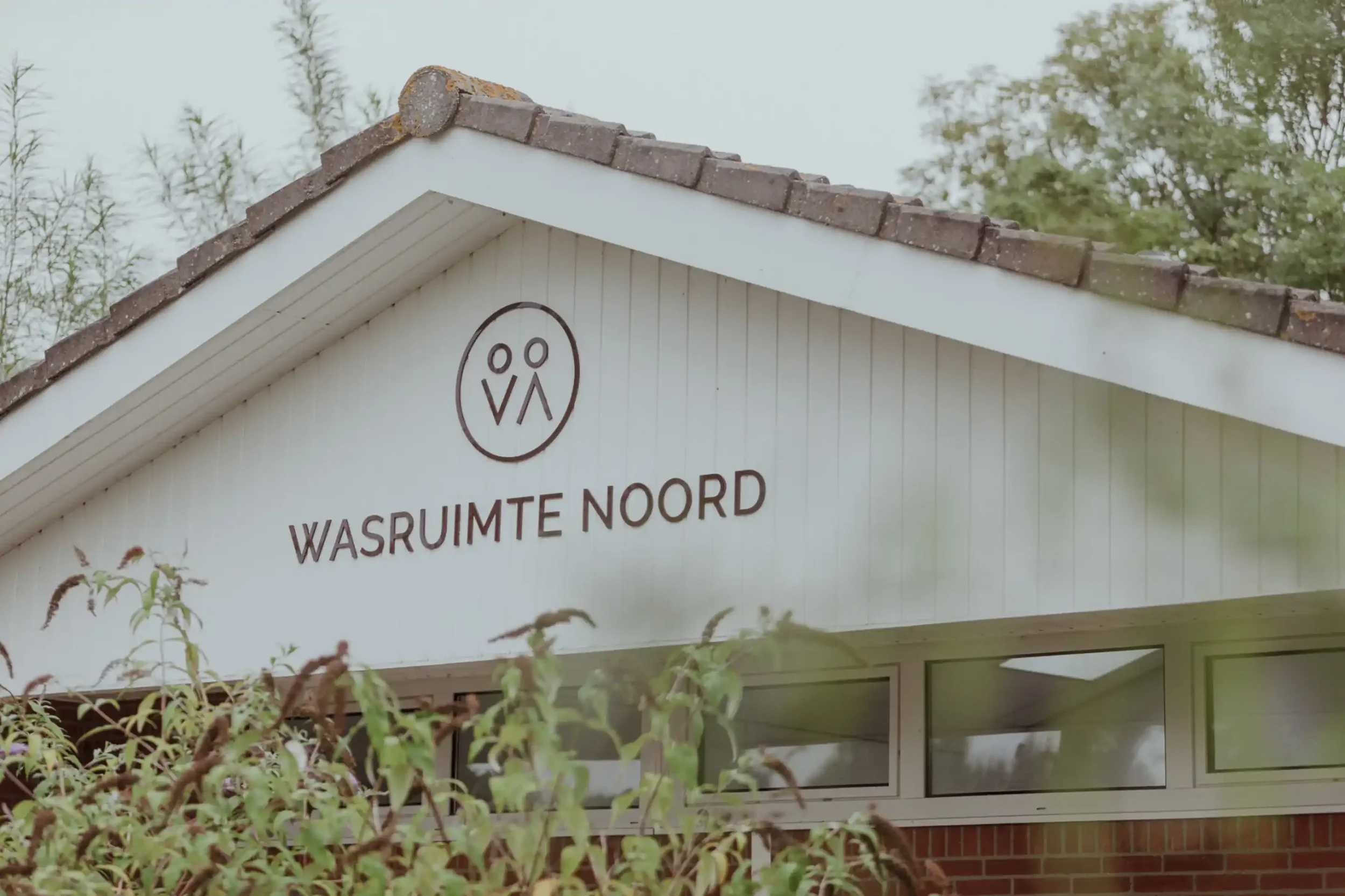 Building with a white exterior and a sloped roof made of dark brown shingles. On the building, there is a logo with a circle and two stylized human figures inside. Below the logo, there is text in both Dutch that reads 'WASRUIMTE NOORD' and 'LAUNDRY ROOM NORTH.'
