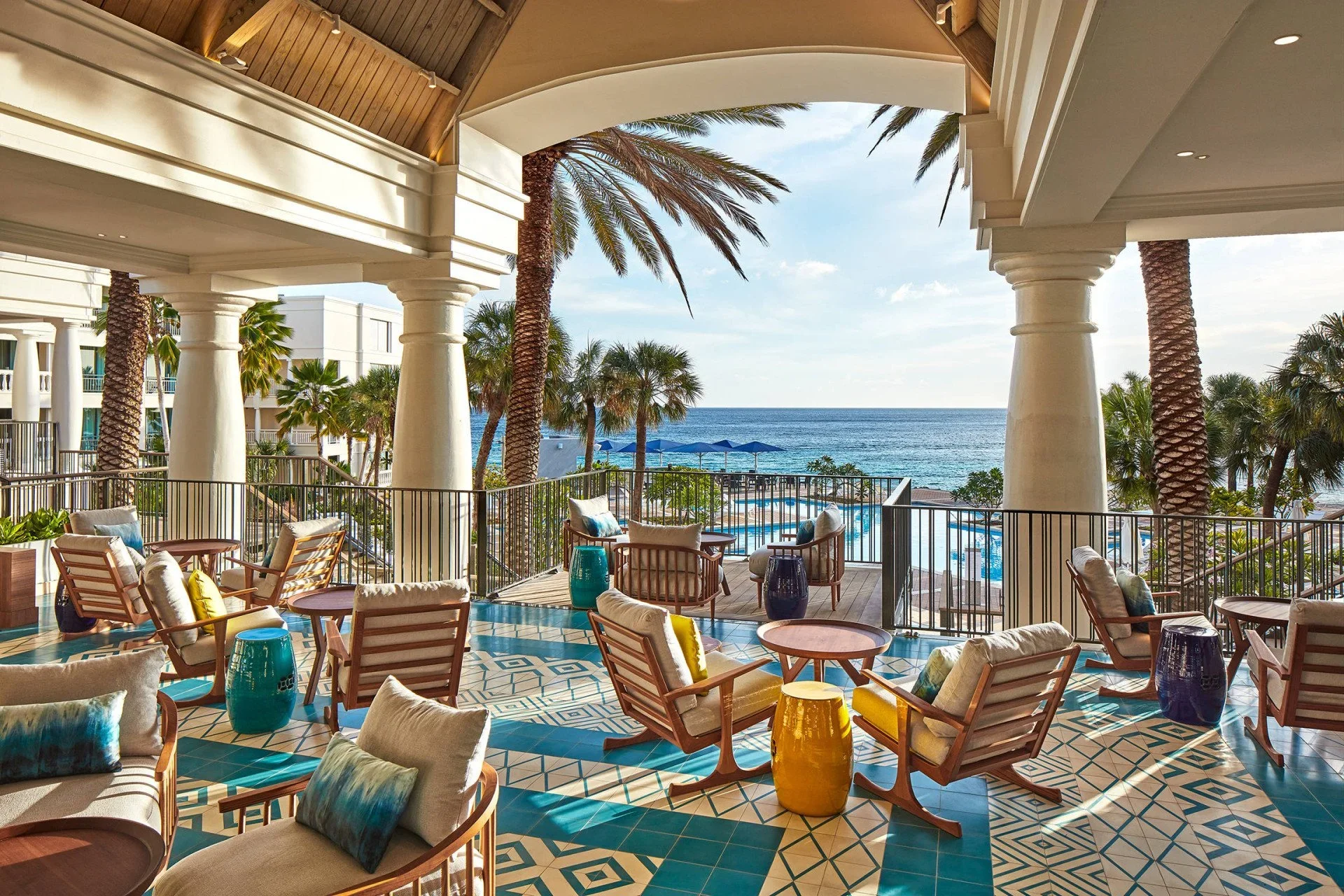 Indoor lounge area of Marriott hotel Curacao with wooden chairs, cushions, and small tables, overlooking a balcony with a view of a pool, palm trees, and the ocean under a partly cloudy sky. BigStoryShort client.