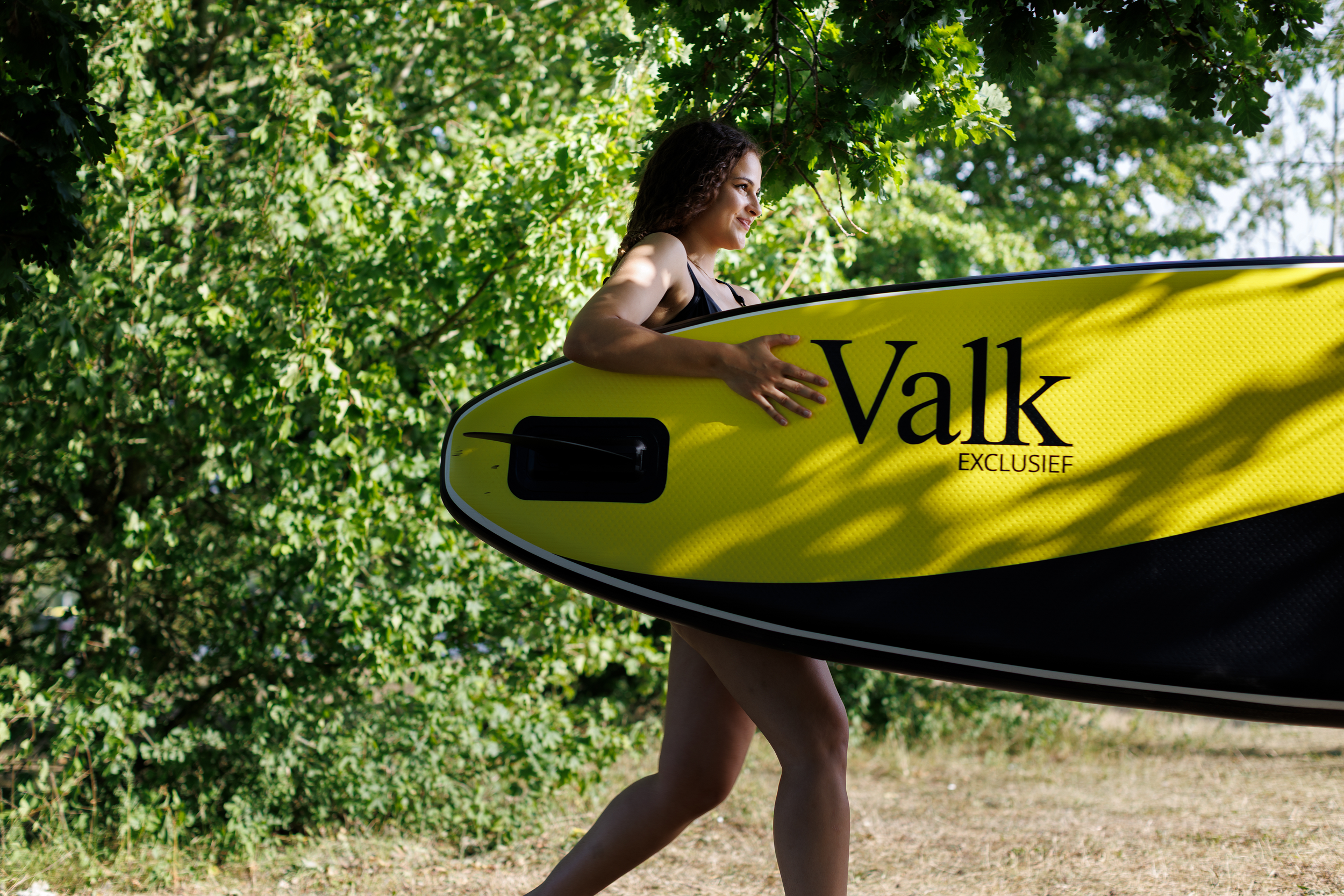 A woman carrying a yellow surfboard with the word 'Van der Valk' written on it, walking outdoors among green trees. BigStoryShort client.