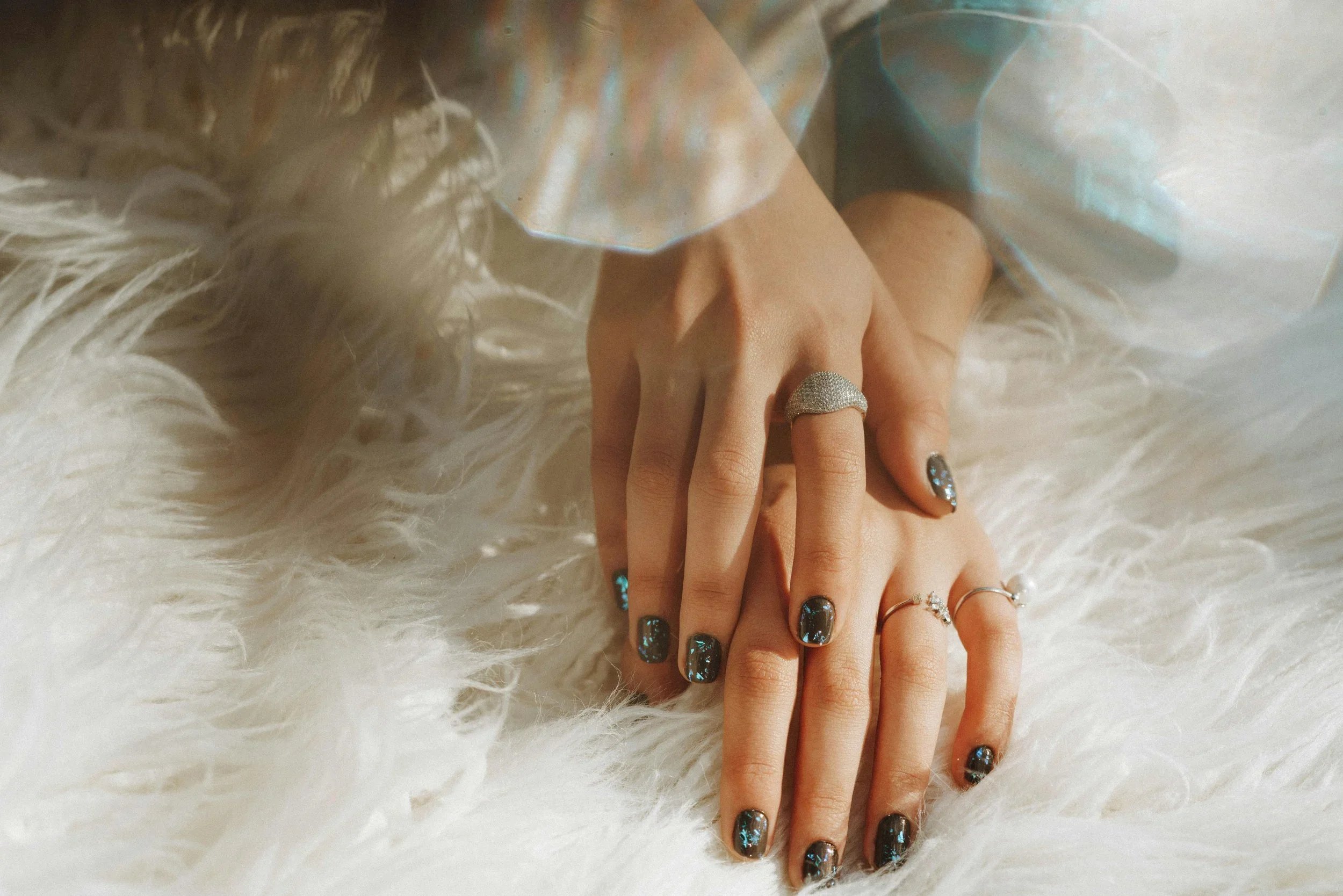 Close-up of hands with black glitter nail polish resting on a white furry surface, adorned with rings from Lucardi Juweliers, with a sheer iridescent fabric and white fluffy material in the background. BigStoryShort client.