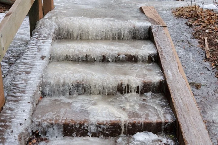 Icy outdoor stairs covered in ice and melting snow, with wooden side rails and wet ground surrounding them.