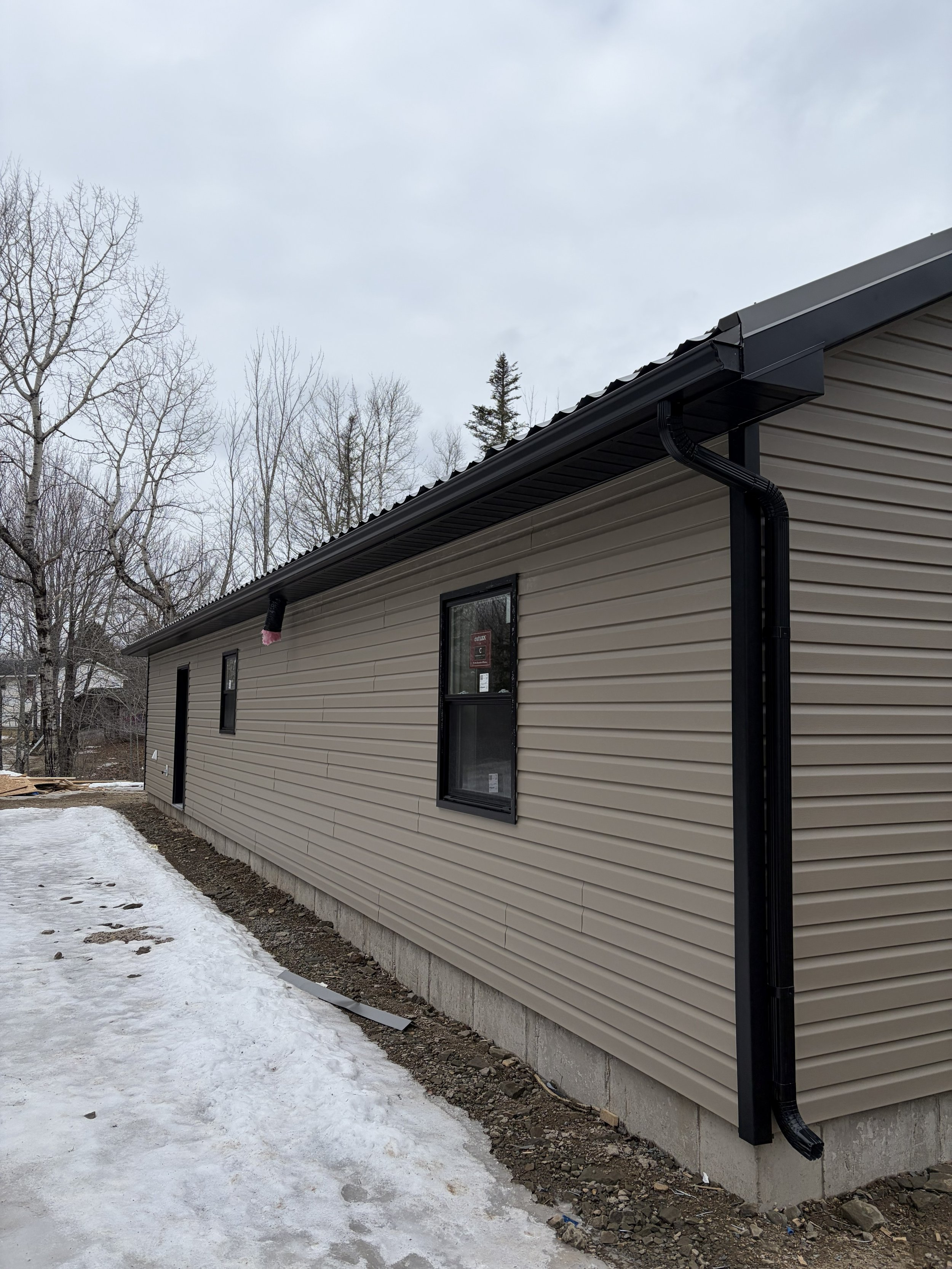 Side view of a beige house with black window frames and a black gutter system, set against a snowy and cloudy sky with bare trees in the background.