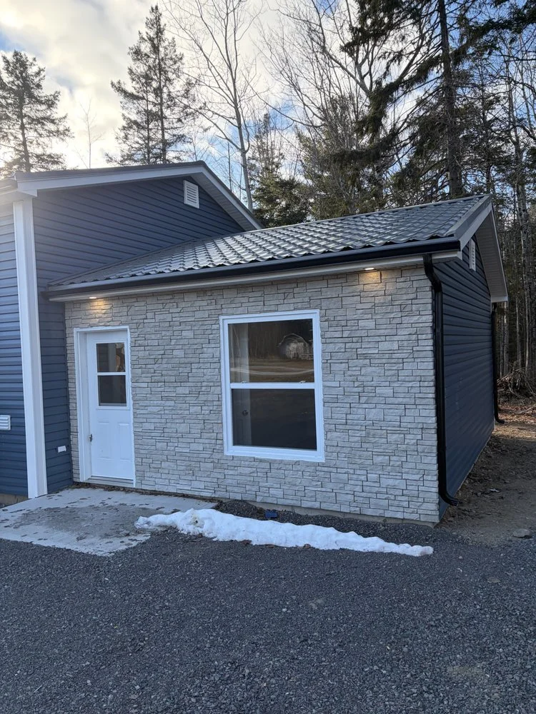 Side of a house with a white door, a window, and exterior lighting, surrounded by trees and a gravel driveway with patches of snow.