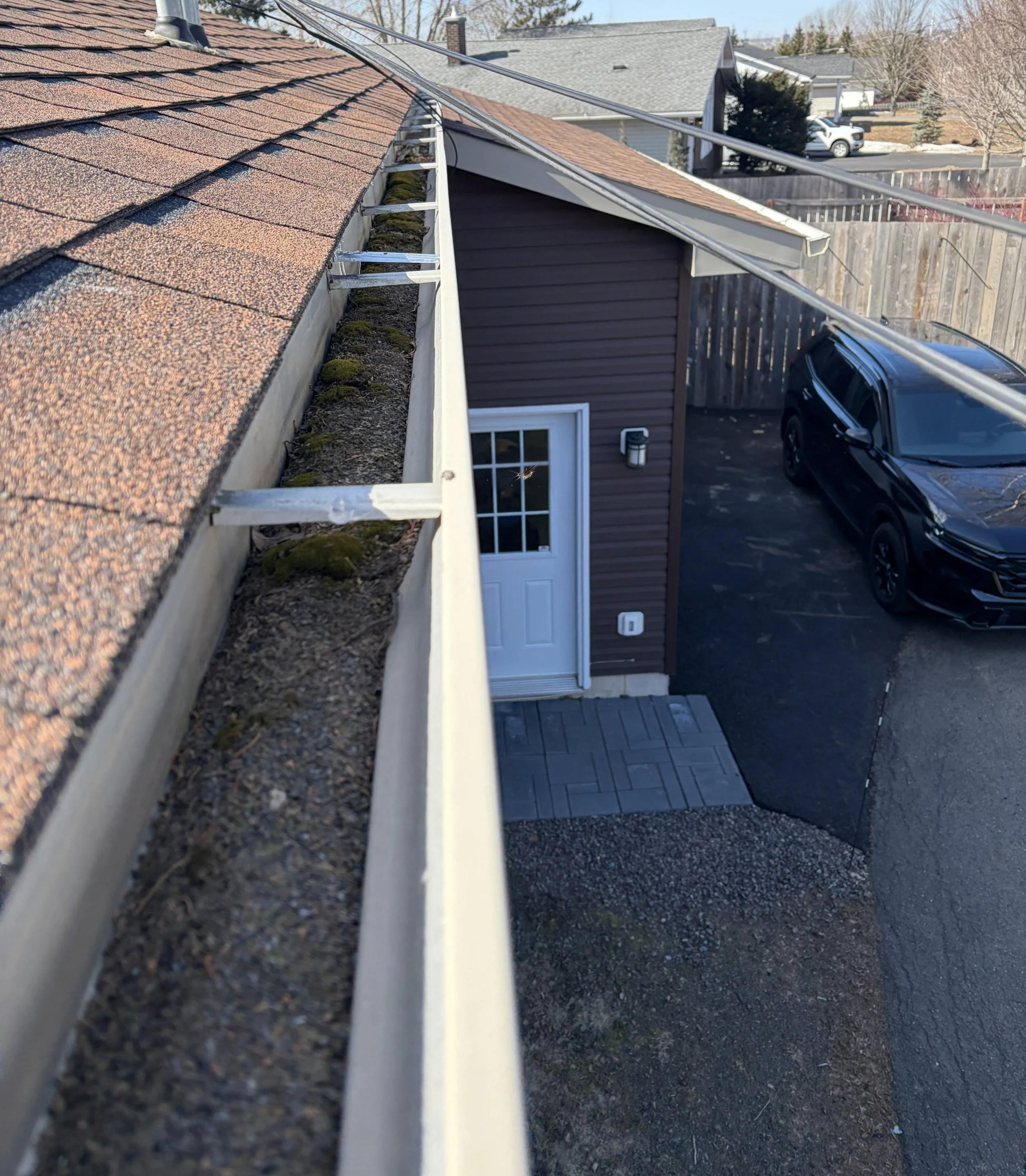 View from the roof showing the shingles, gutter, moss, and snow on the ground outside a residential house with a driveway and black car.