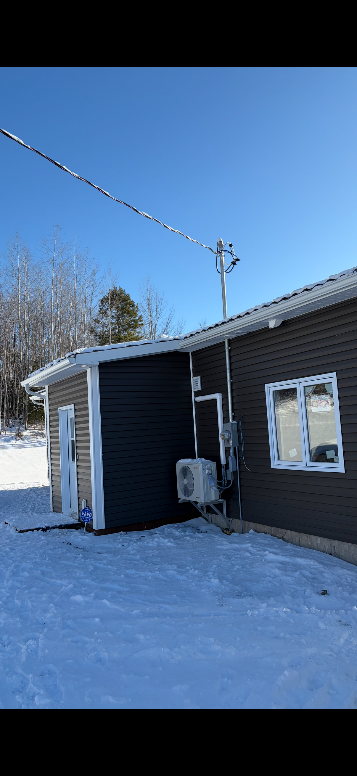 A house exterior in winter with snow on the ground, a black house siding, a white-framed window and door, an outdoor air conditioning unit, and a utility pole with power lines against a clear blue sky.