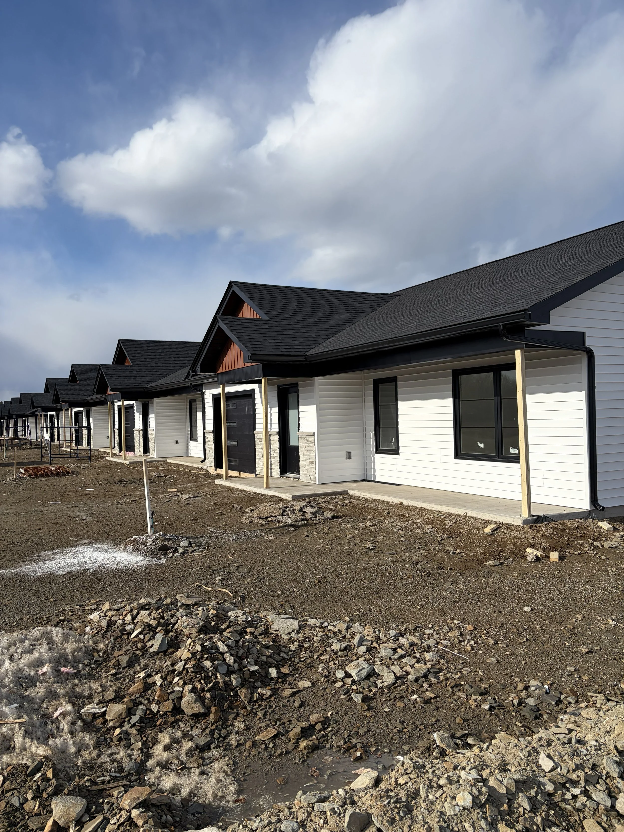 Newly constructed row of houses with black roofs and white siding, under construction, with dirt and gravel in the foreground and a partly cloudy sky above.