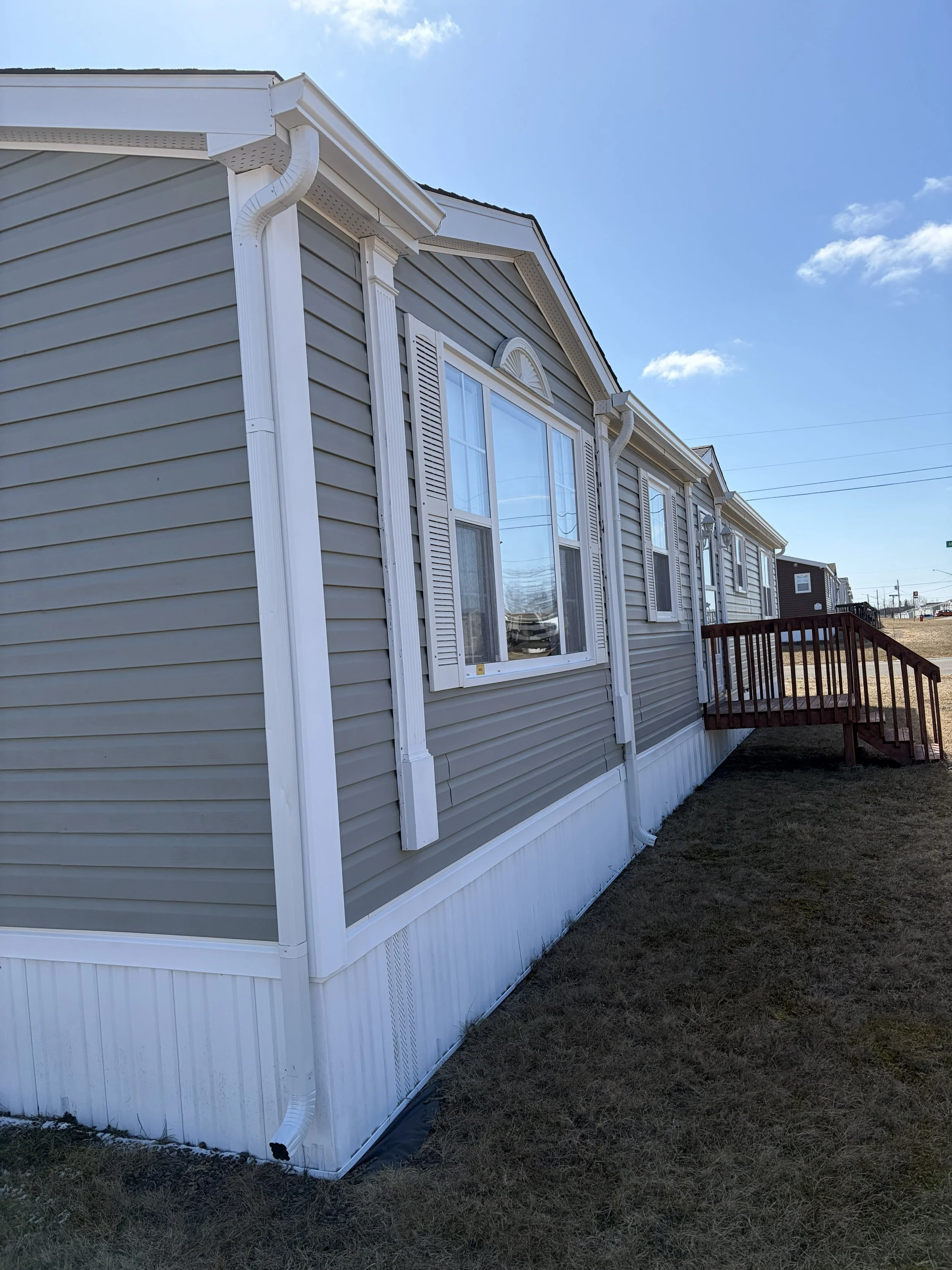 Side view of a modern manufactured home with gray siding, white trim, and a red staircase leading to an entrance, set on a grassy lot under a blue sky.