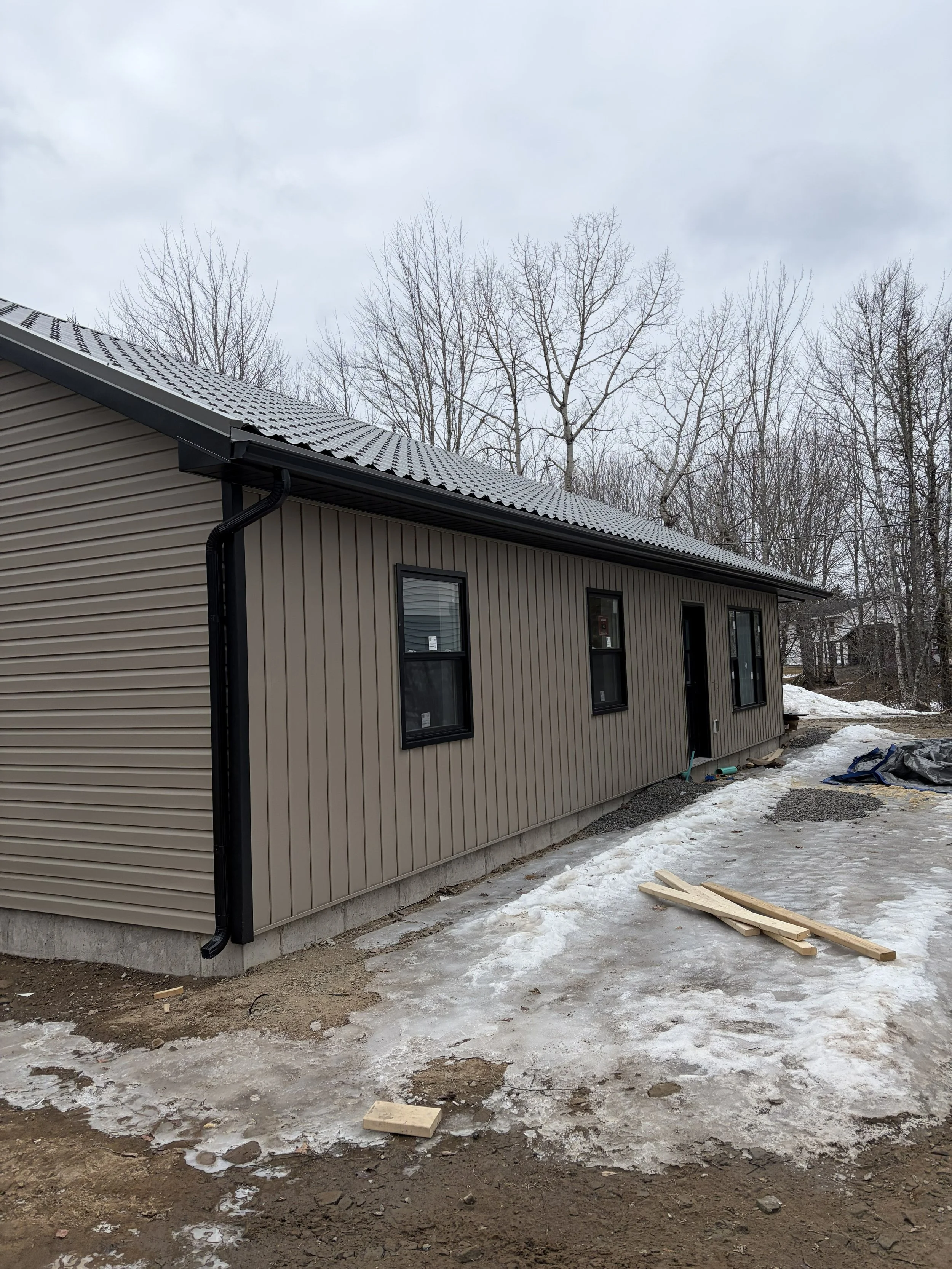 Exterior view of a new house under construction with beige siding, black window frames, and a metal roof, with snow and ice on the ground and some wooden planks in the yard.