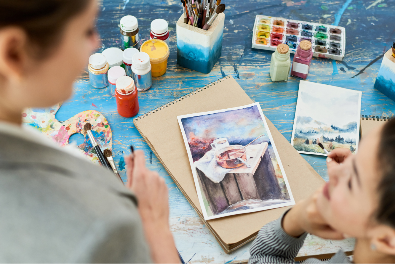 Two children working on watercolor paintings at a table with art supplies, including jars of paint, brushes, a watercolor palette, and watercolor paintings.