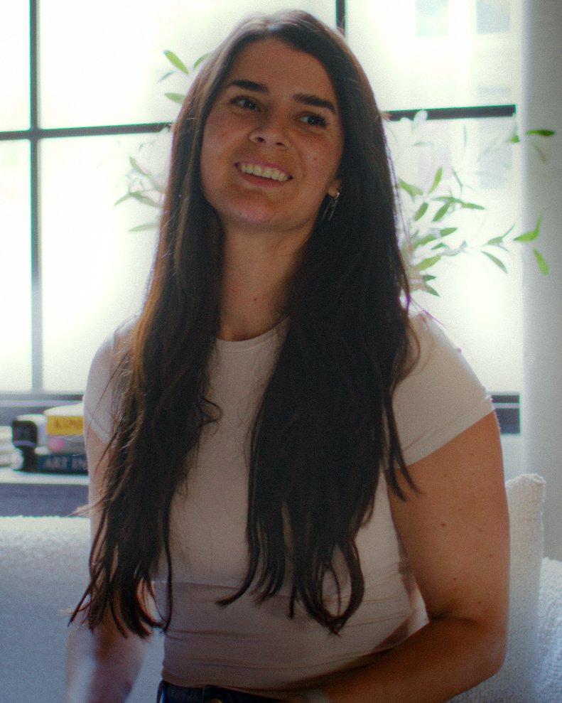 A woman with long dark hair smiling indoors near a window with green plants in the background.