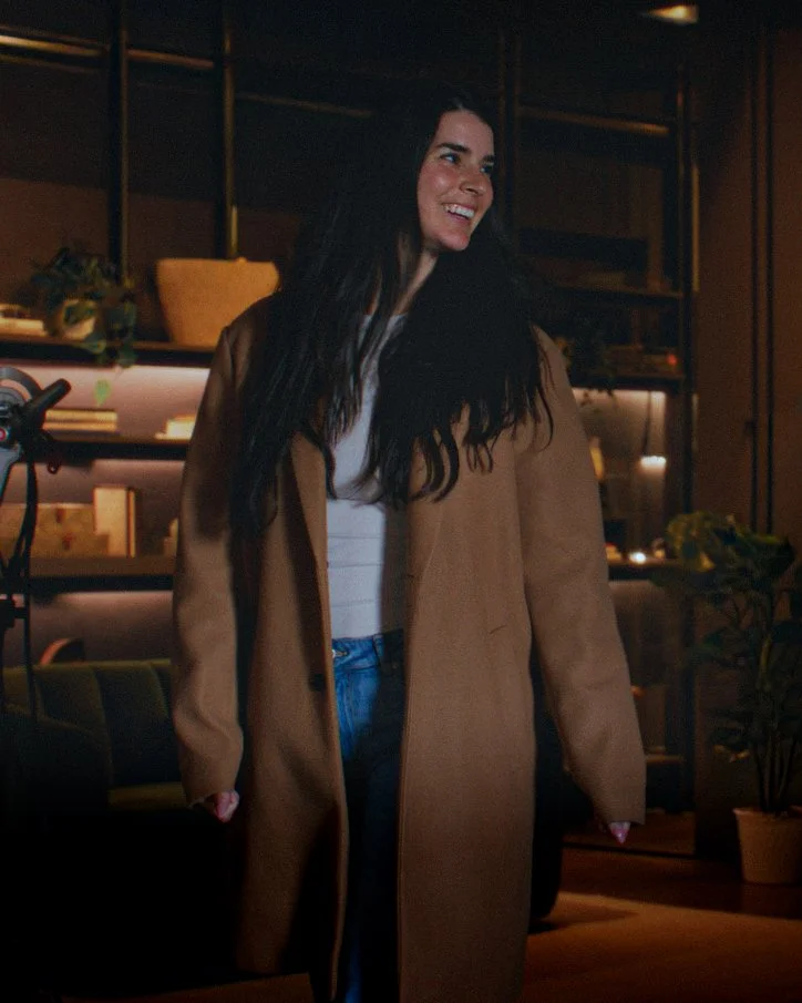 A woman with long dark hair, wearing a tan coat over a white shirt with blue jeans, standing in a warmly lit room with modern decor and plants, smiling.