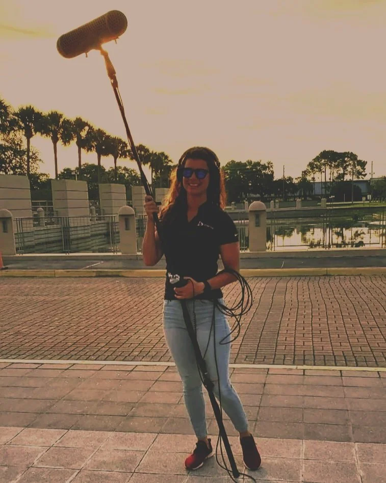 Woman holding a boom microphone outdoors at sunset, with palm trees and water in the background.