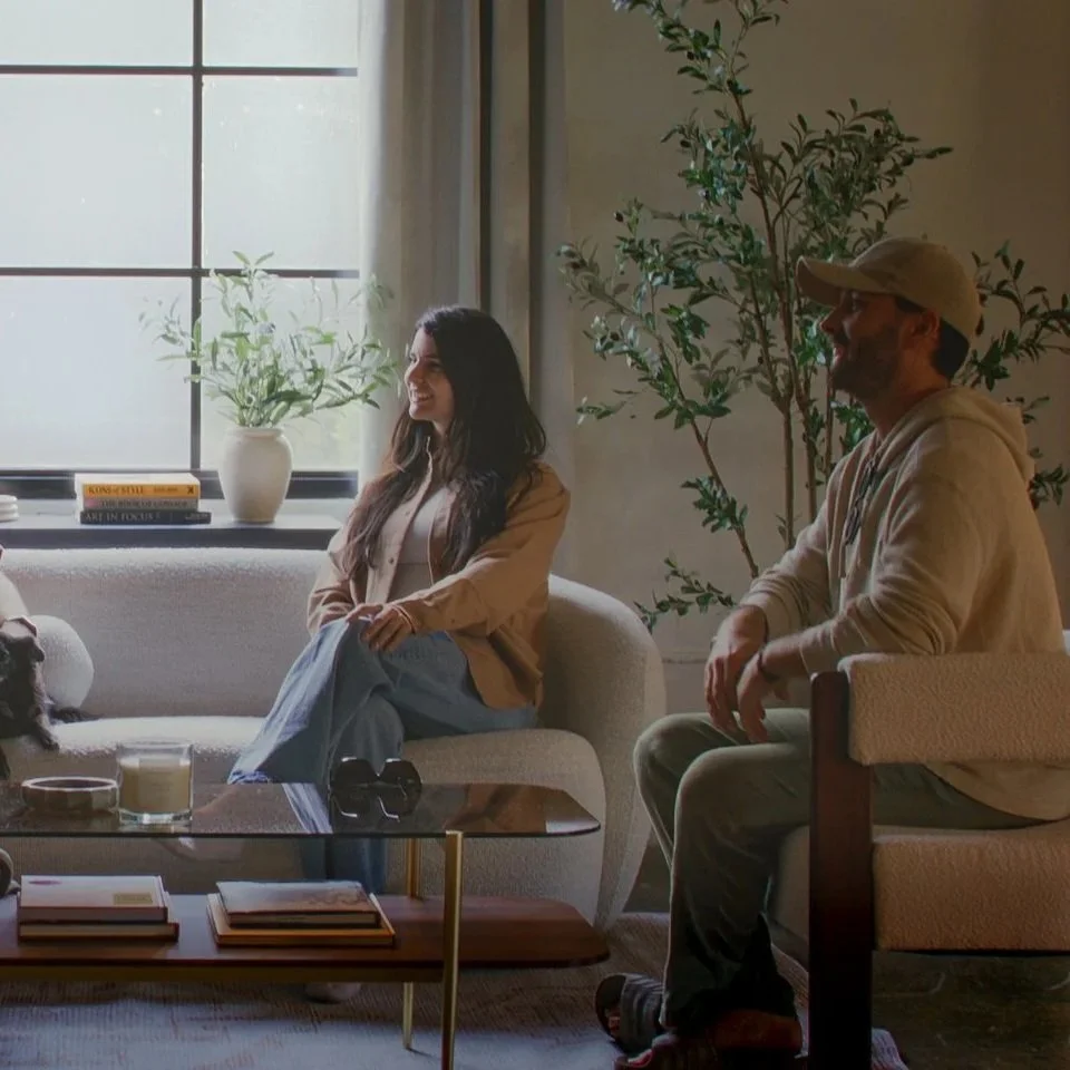 A woman with long dark hair and a man with a baseball cap sitting in a cozy living room, engaged in a conversation. They are near a window with natural light, a potted plant, and books on the window sill.