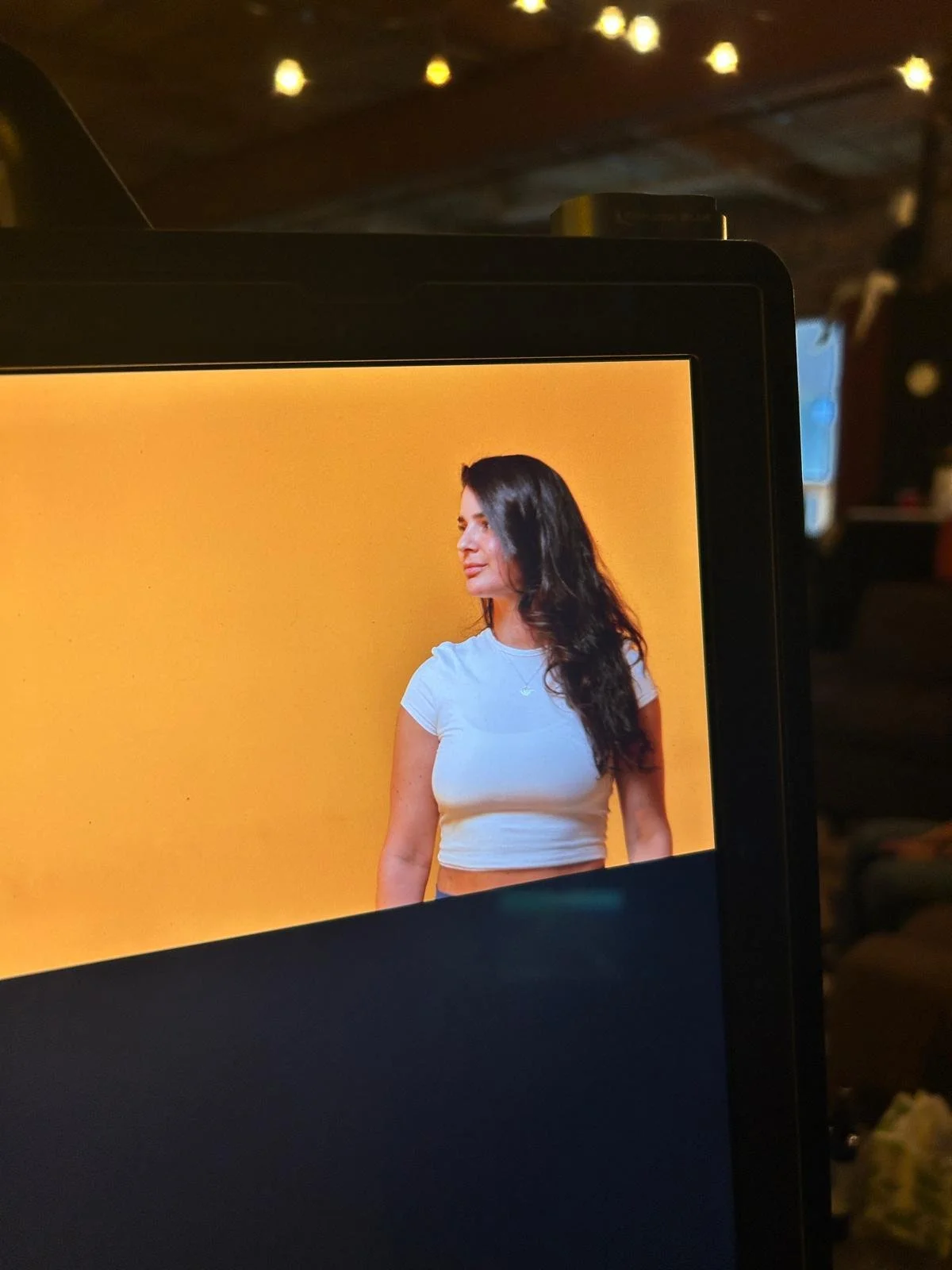 A woman with long dark hair wearing a white crop top, standing against a yellow background, is seen on a computer monitor.