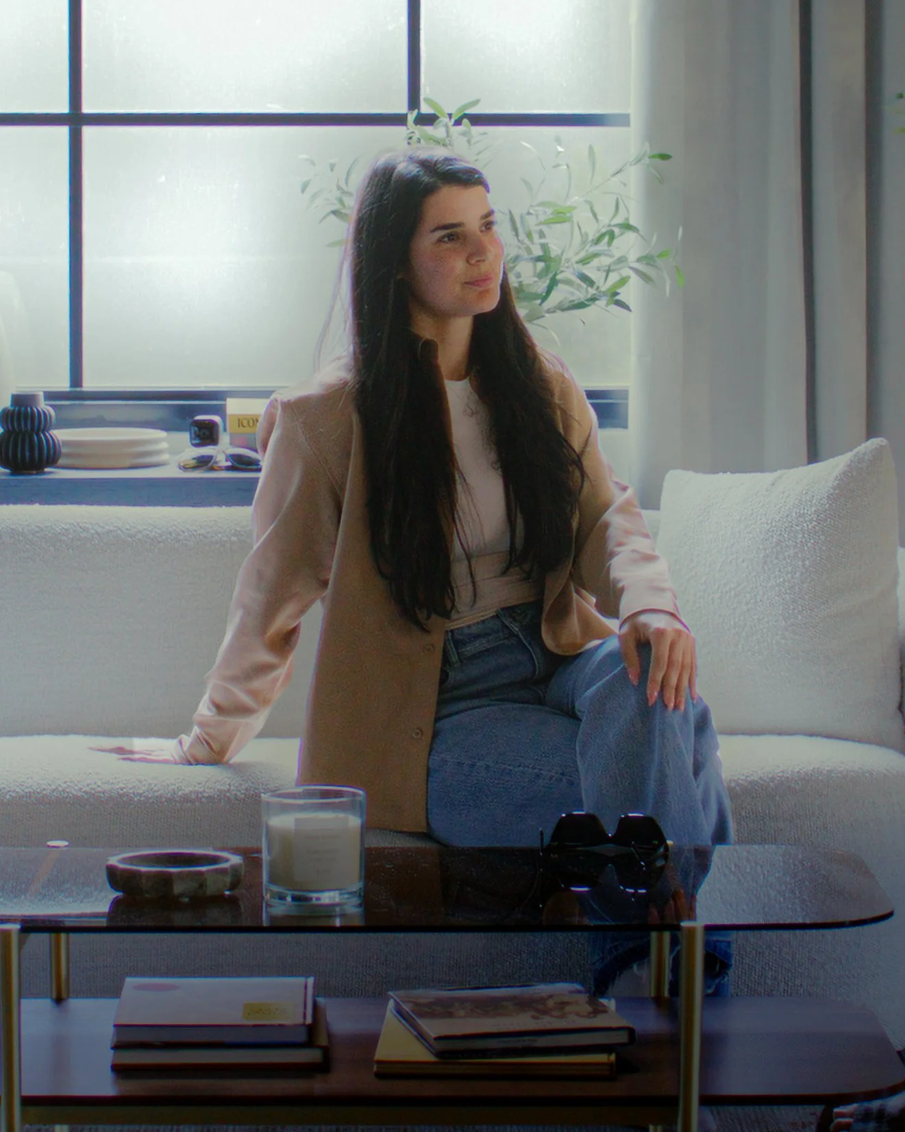 A woman sitting on a cream-colored sofa in front of a window, with a plant behind her, wearing a light brown blazer and blue jeans, with books and glasses on a coffee table in front of her.