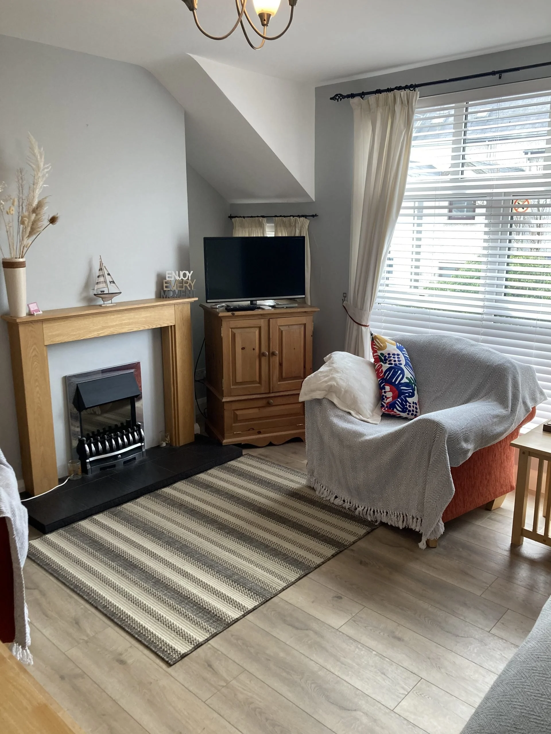 Living room with a fireplace, a wooden TV stand with a flat-screen TV, a large window with blinds and curtains, an armchair with a blanket and decorative pillow, a striped rug, and a partial view of another chair.