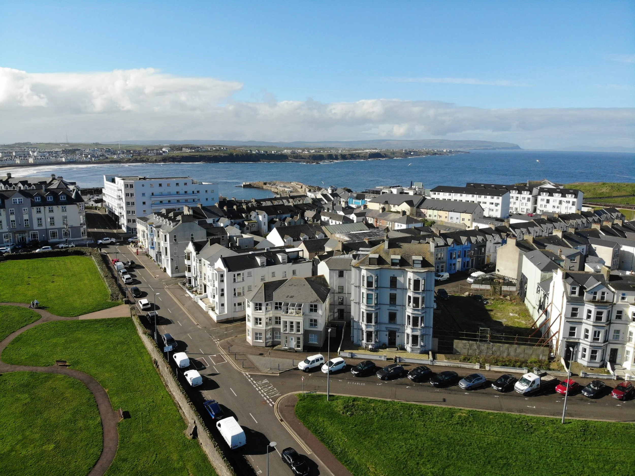 A coastal town with white multi-story buildings, green park, parking lot, and ocean in the background under blue sky with clouds.