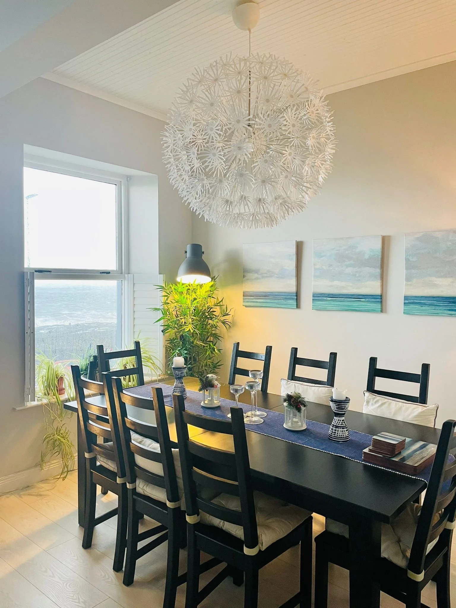 A dining room with a black wooden table and eight matching chairs with cream-colored cushions. The table is decorated with a navy table runner, candles, and small floral arrangements. A large, white, spherical, spiky pendant light hangs from the ceil