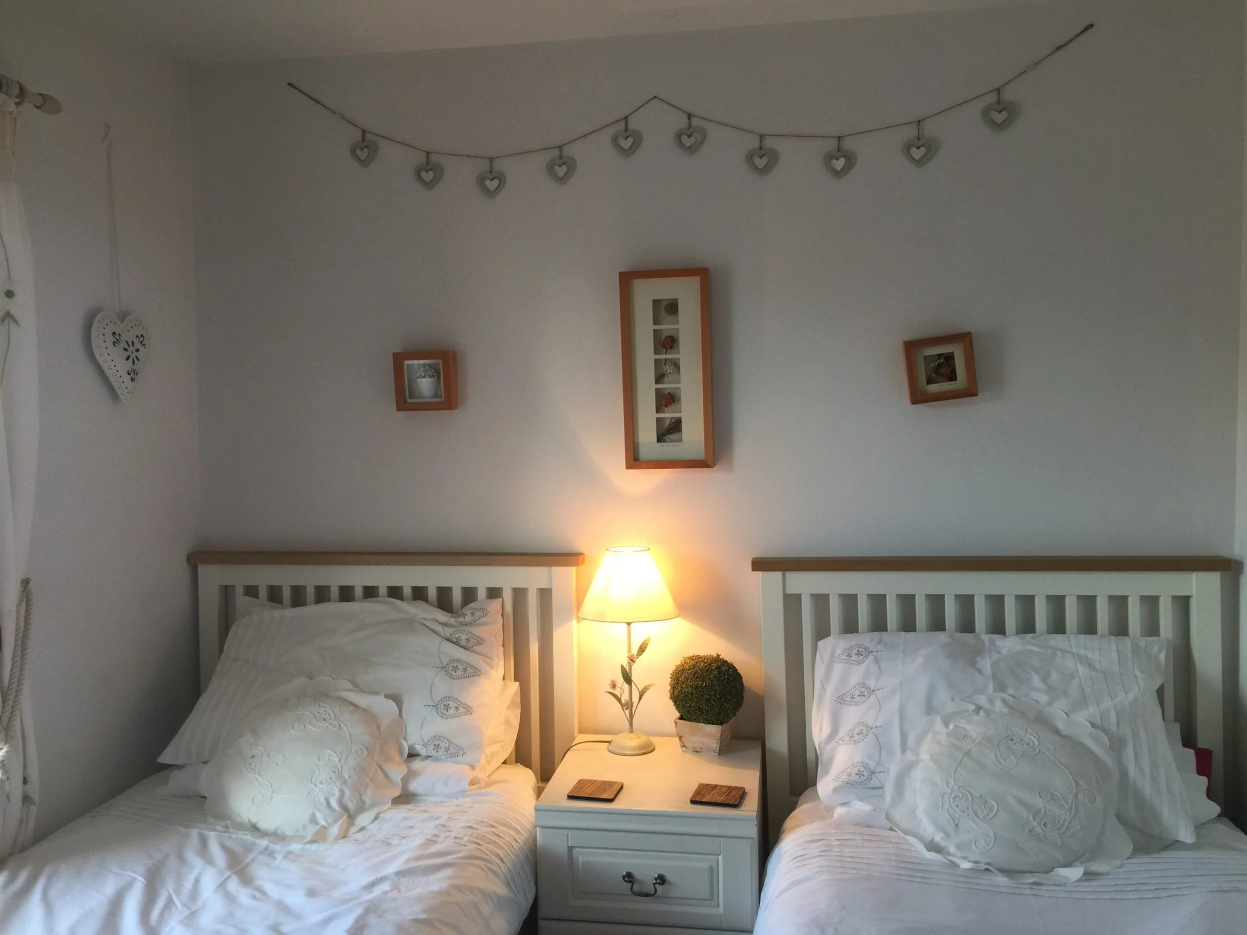 A bedroom with two white beds, a window with white curtains, a nightstand with a lamp and decorative items, and heart-shaped wall decor.
