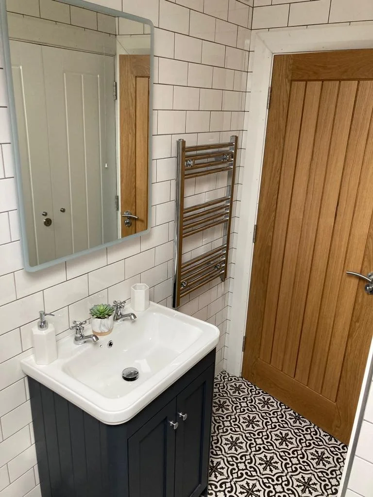 Small bathroom with white tiled walls, wooden door, rectangular mirror, black vanity with white sink, small potted plant, soap dispensers, chrome towel rack, and patterned black and white floor tiles.