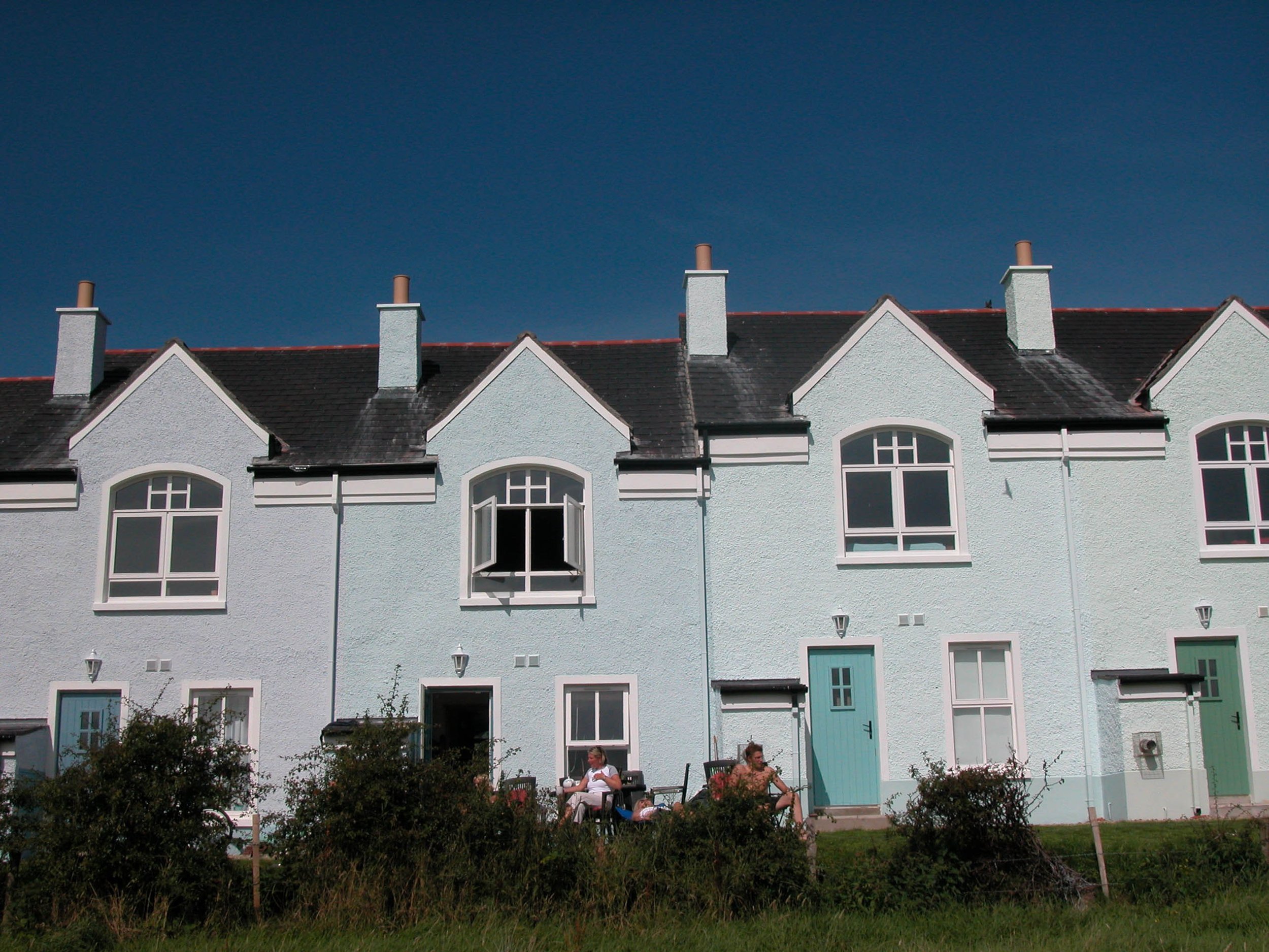 Row of white seaside houses with blue doors and windows, some with open shutters, under a clear blue sky, with two women sitting outside in front of the houses.