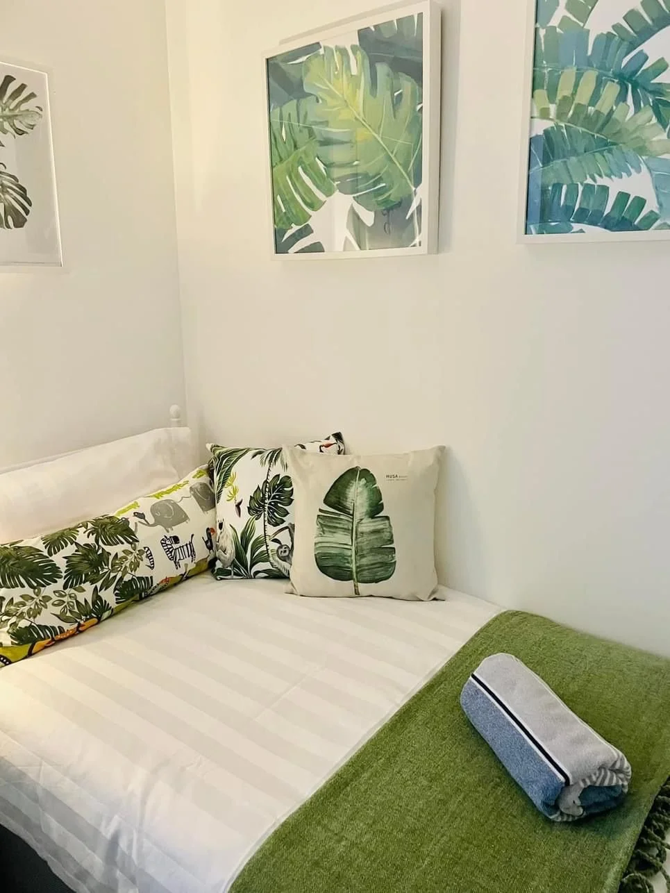 A neatly made bed with tropical-themed pillows, a rolled towel on a green bedcover, and framed botanical artwork on the white wall.